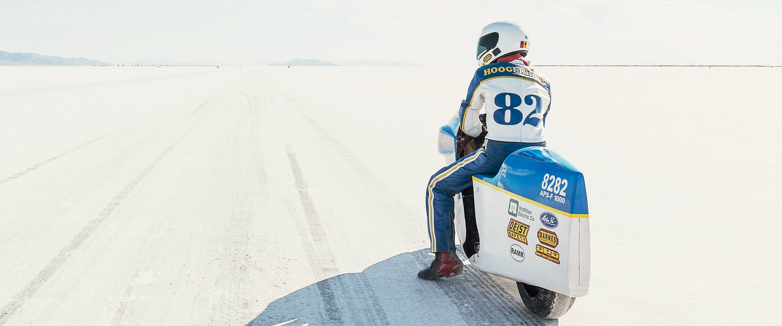 A land speed motorcycle racer wearing a blue and yellow race suit numbered 82 sits astride a white streamlined motorcycle numbered 8282 on the Bonneville Salt Flats, facing away from the camera toward the long straight track that stretches to the distant mountains and pale horizon. Tyre tracks score the white salt surface at the start line as a soft blue sky with scattered clouds completes the vast open scene. Fine art colour photographic print by Simon Davidson.
