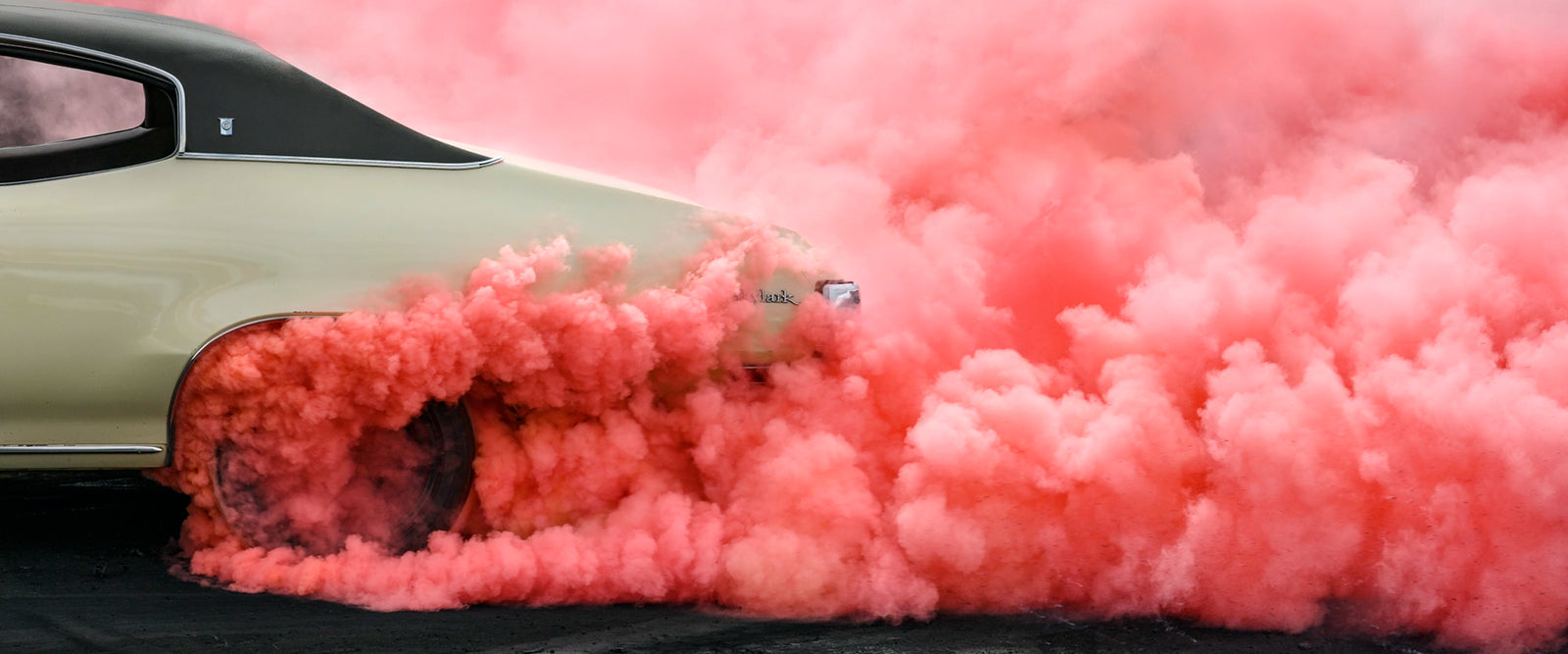 Rear side quarter of a metallic gold Buick SKYLARK, with black vinyl roof performs a burnout, engulfed in a dense rolling cloud of vivid watermelon coloured tyre smoke that billows across the entire right side of the frame. The smoke erupts from beneath the rear wheel arch and sweeps dramatically across the scorched black bitumen, the gold body and dark vinyl roof barely visible through the churning watermelon haze. Fine art colour photographic print by Simon Davidson.