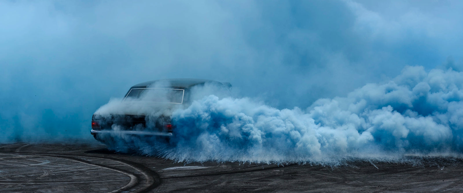 A black Holden sedan known as UCSMOKE performing a burnout, almost completely dissolved into a vast rolling cloud of deep blue tyre smoke. Fine art burnout photographic print by Simon Davidson.