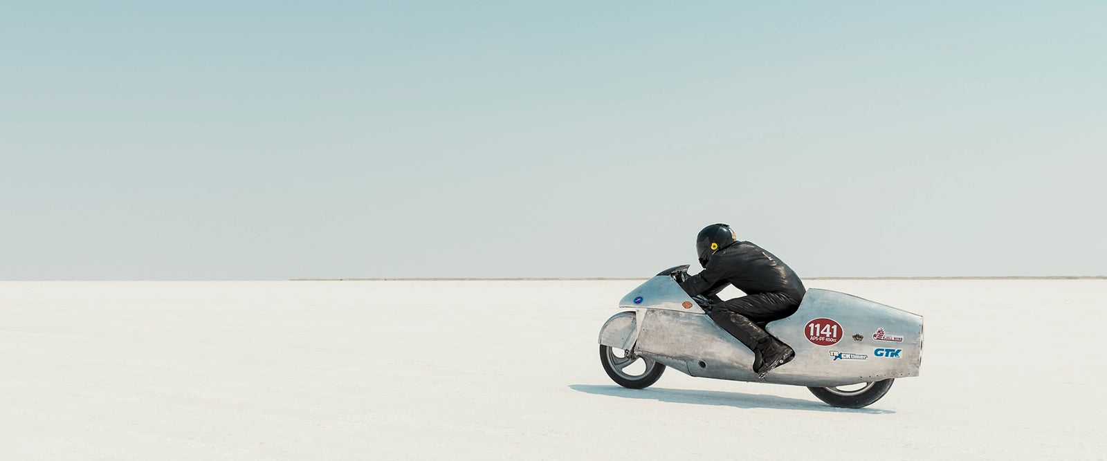 Clad in black leather, a rider crouches low over a bare metal aluminium streamliner motorcycle numbered 1141 as it races across the vast white expanse of the bonneville salt flats The rider and machine are small but in striking form against the pale horizon and soft blue sky above. The hand beaten metal bike and minimal silhouette give the scene a timeless, sculptural quality as it pushes across the featureless white surface. Fine art colour photographic print by Simon Davidson.