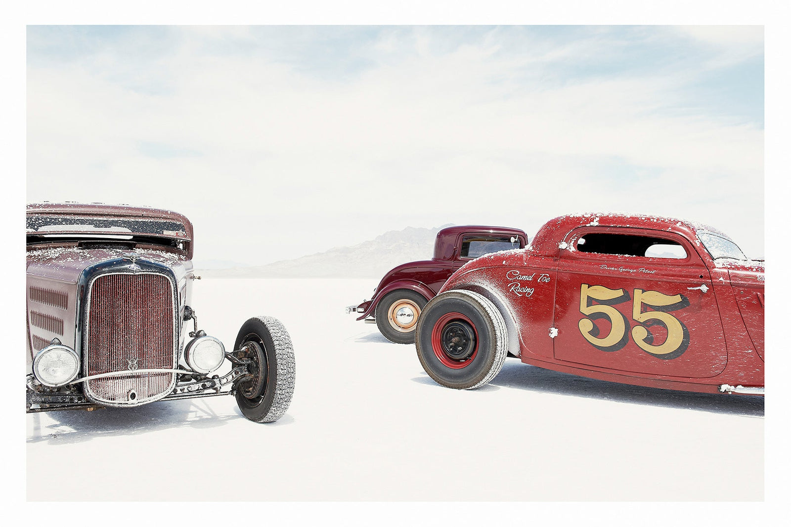 Two salt encrusted Rolling Bones hot rods sit on the blinding white surface of the Bonneville Salt Flats, their weathered bodies dusted with salt crystal residue. A third maroon coupe visible between them in the middle distance. A pale mountain range dissolves into the white horizon behind them. Fine art colour photographic print by Simon Davidson.