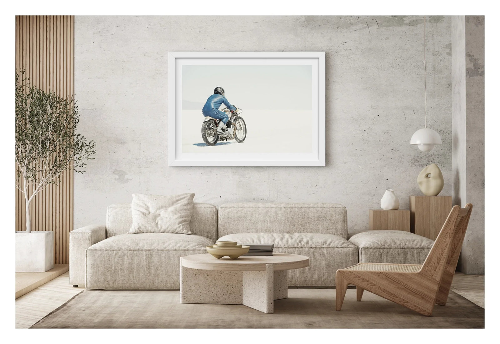 Framed in white. A lone motorcycle racer dressed in a blue leather suit and silver helmet rides a vintage Triumph, land speed racing motorcycle across the blinding white expanse of the Bonneville salt flats, the rider and bike reduced to a small solitary figure against an almost entirely white horizon and washed out sky. Shown in a contemporary living room. Fine art colour photographic print by Simon Davidson.