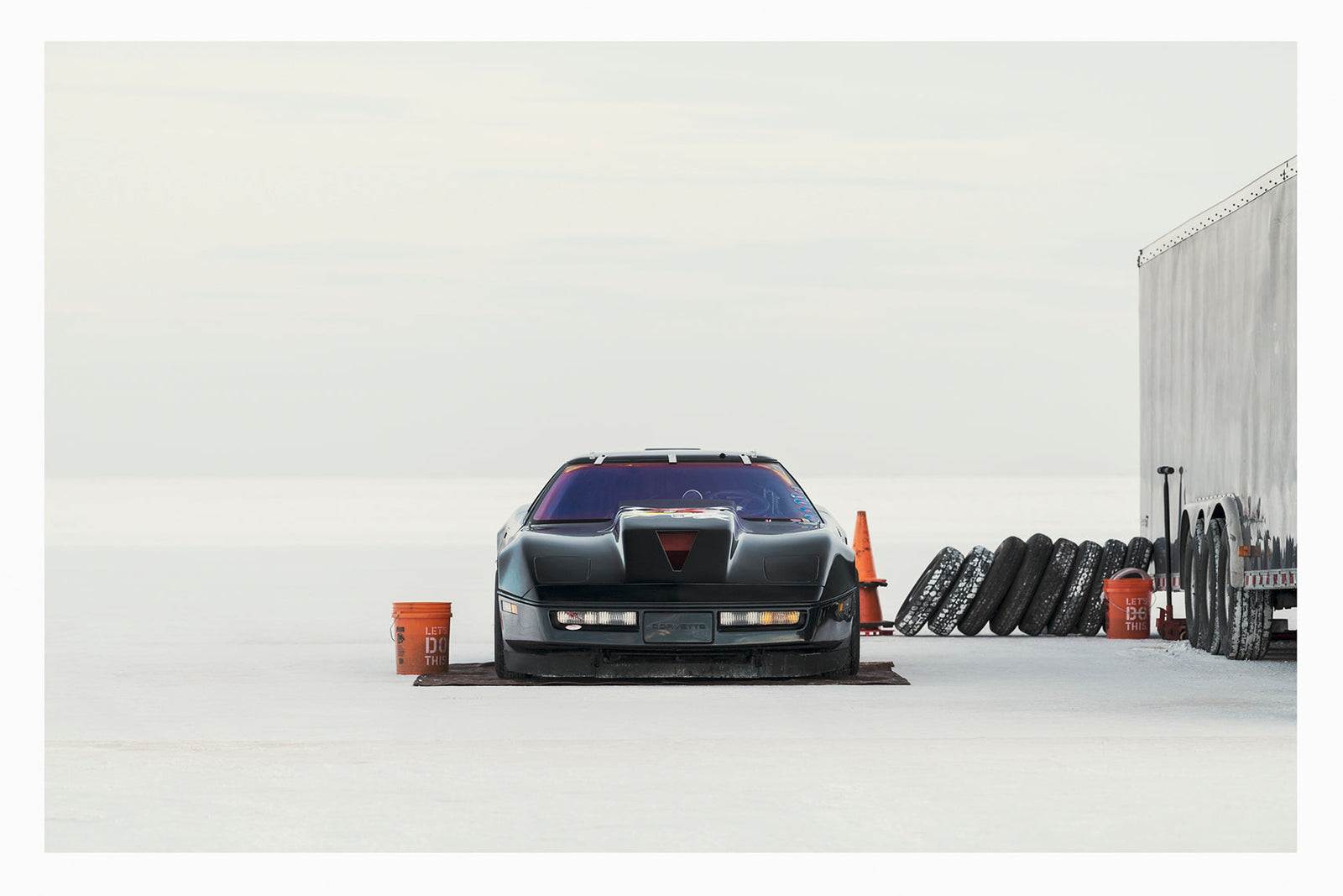 A black Chevrolet Corvette land speed racing car sits low on a ground sheet in the pits at Bonneville. Surrounded by the vast white expanse of the Salt Flats, faced directly toward the camera. Orange buckets, a row of spare tyres leaning against a trailer and an orange traffic cone are scattered around the pit area to the right, the whole scene dissolving into a featureless white horizon and sky. Fine art colour photographic print by Simon Davidson.