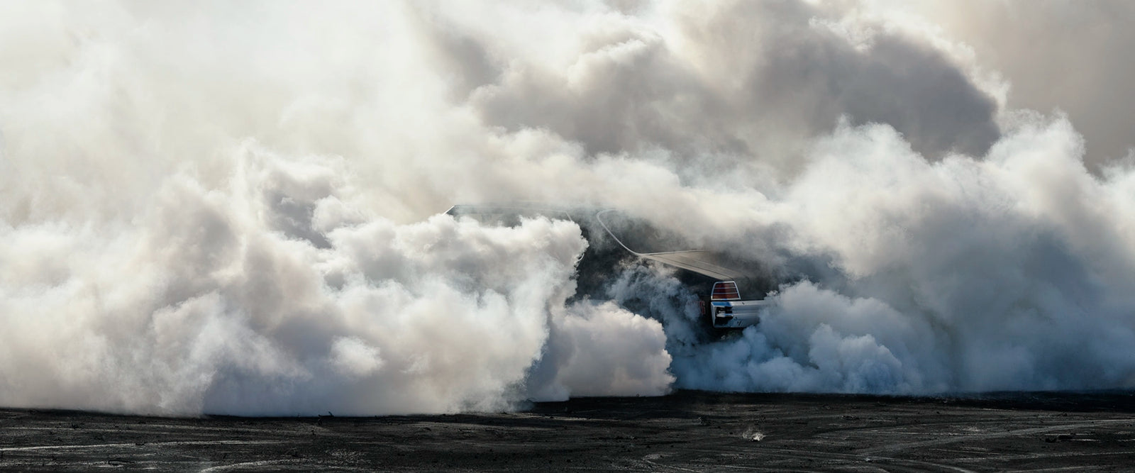 We see the rear of a black Holden GTS sedan is almost entirely swallowed by a towering cloud of white tyre smoke during a burnout, with only the red tail light and rear chrome bumper visible through the billowing haze. The smoke builds into a massive marshmallow like formation that fills the frame from the black bitumen covered in burnout detritus to the pale sky above, dwarfing the car beneath it. Fine art colour photographic print by Simon Davidson.