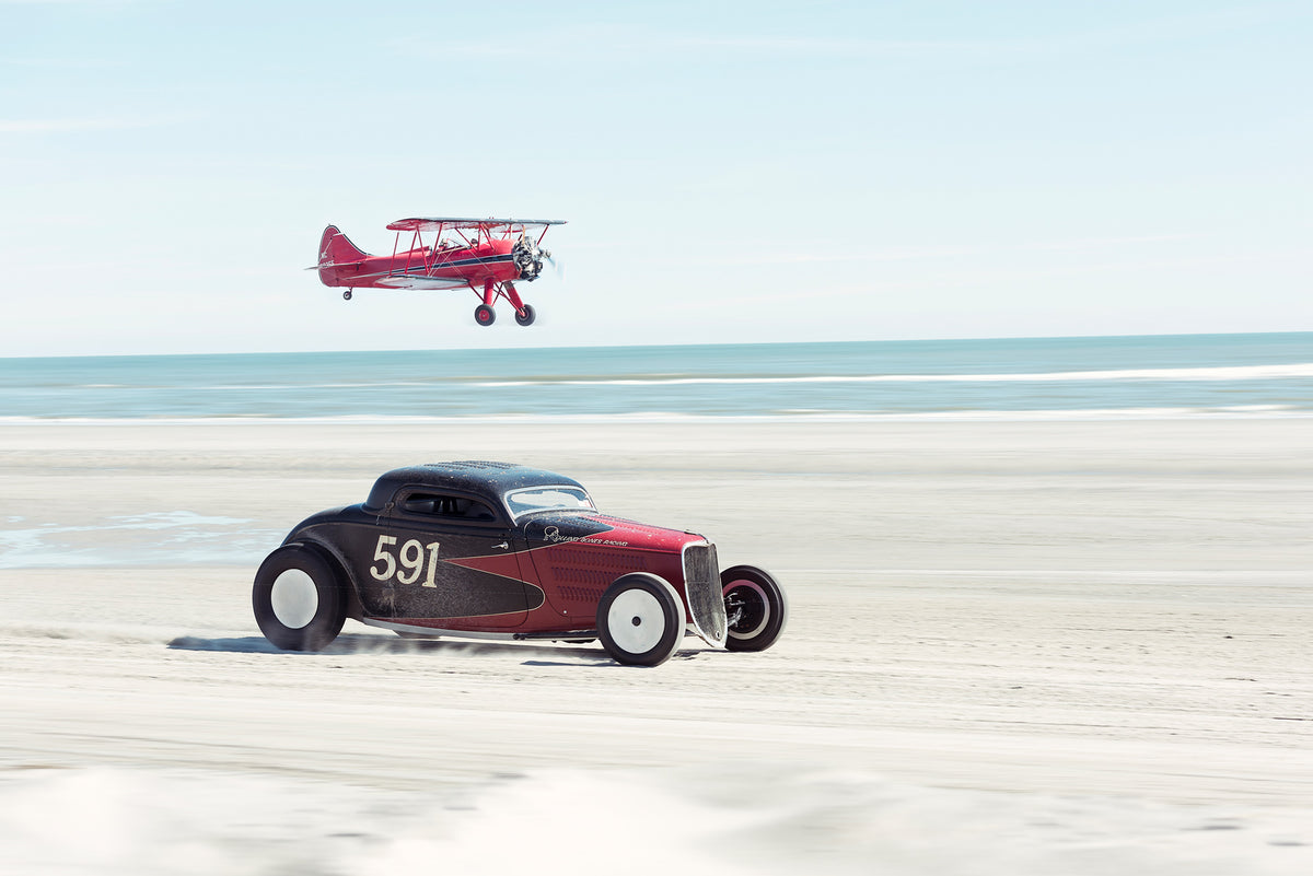 Ken Schmidt driving his red and black custom hot rod coupe from Rolling Bones. Numbered 591, racing along the beach while a red biplane flies low overhead, the ocean stretching out behind them. Captured at the Race of Gentlemen. Fine art colour photographic print by Simon Davidson.