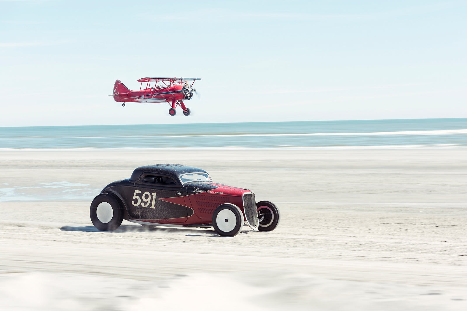 Ken Schmidt driving his red and black custom hot rod coupe from Rolling Bones. Numbered 591, racing along the beach while a red biplane flies low overhead, the ocean stretching out behind them. Captured at the Race of Gentlemen. Fine art colour photographic print by Simon Davidson.