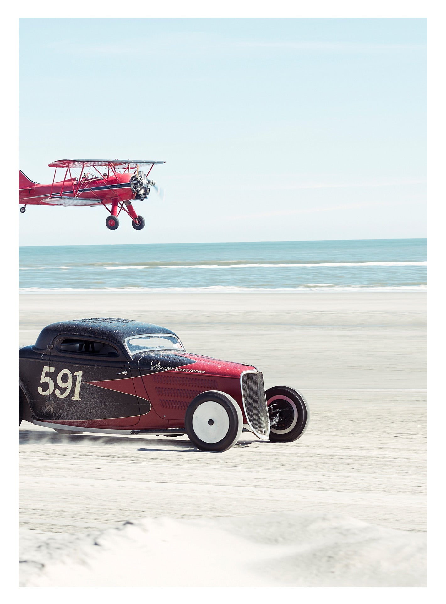 Ken Schmidt driving his red and black custom hot rod coupe from Rolling Bones. Numbered 591, racing along the beach while a red biplane flies low overhead, the ocean stretching out behind them. Captured at the Race of Gentlemen. Fine art colour photographic print by Simon Davidson.