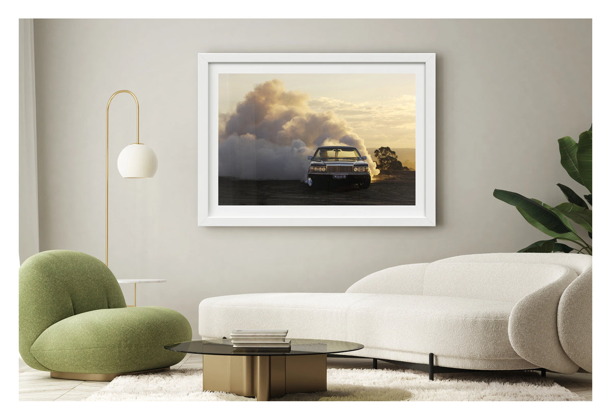 Framed in white. Front view of a WB Holden ute performing a poetic burnout at golden hour, smoke billowing behind it like an approaching thunderstorm. Fading sunlight catches the haze and chrome as the car stays ahead of its own chaos. Shown in a contemporary living space. Fine art print by Simon Davidson.