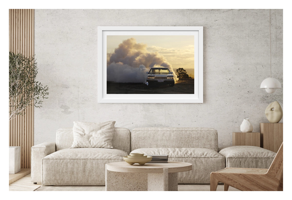 Framed in white. Front view of a WB Holden ute performing a poetic burnout at golden hour, smoke billowing behind it like an approaching thunderstorm. Fading sunlight catches the haze and chrome as the car stays ahead of its own chaos. Shown in a contemporary living space. Fine art print by Simon Davidson.