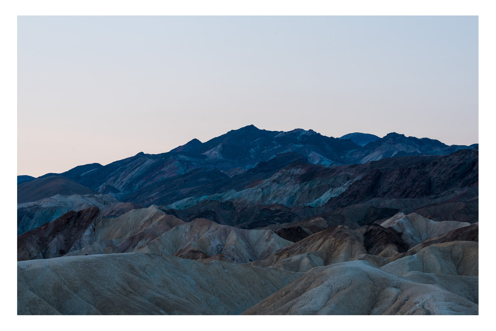 The layered badlands of Zabriskie Point in Death Valley at first light, the eroded mudstone formations in shades of tawny gold and grey in the foreground, the Black Mountains rising in deep blue and teal behind them under a pale pink sky. Fine art colour photographic print by Simon Davidson.