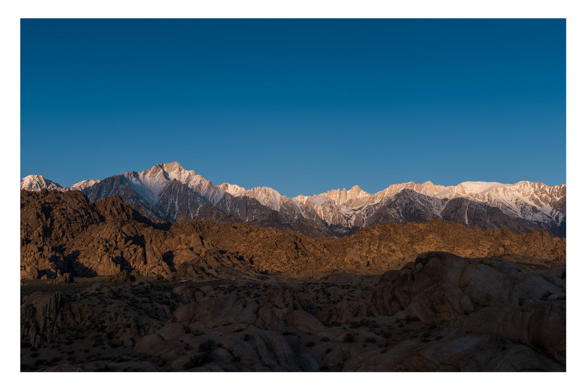 The granite boulders of the Alabama Hills glowing warm in early morning light with the snow capped Sierra Nevada including Mount Whitney rising sharply behind them against a clear blue sky. Fine art colour photographic print by Simon Davidson.