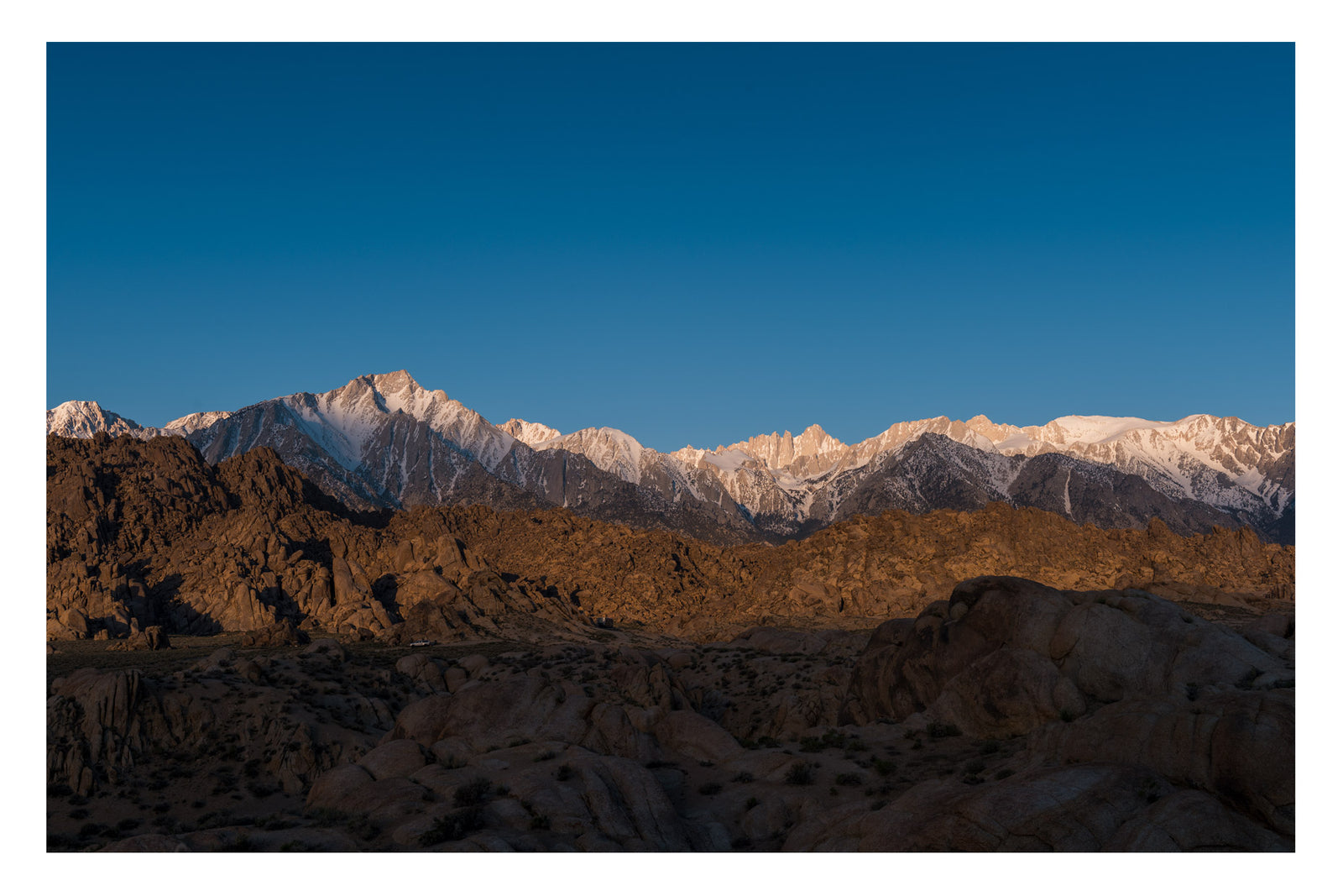 The granite boulders of the Alabama Hills glowing warm in early morning light with the snow capped Sierra Nevada including Mount Whitney rising sharply behind them against a clear blue sky. Fine art colour photographic print by Simon Davidson.