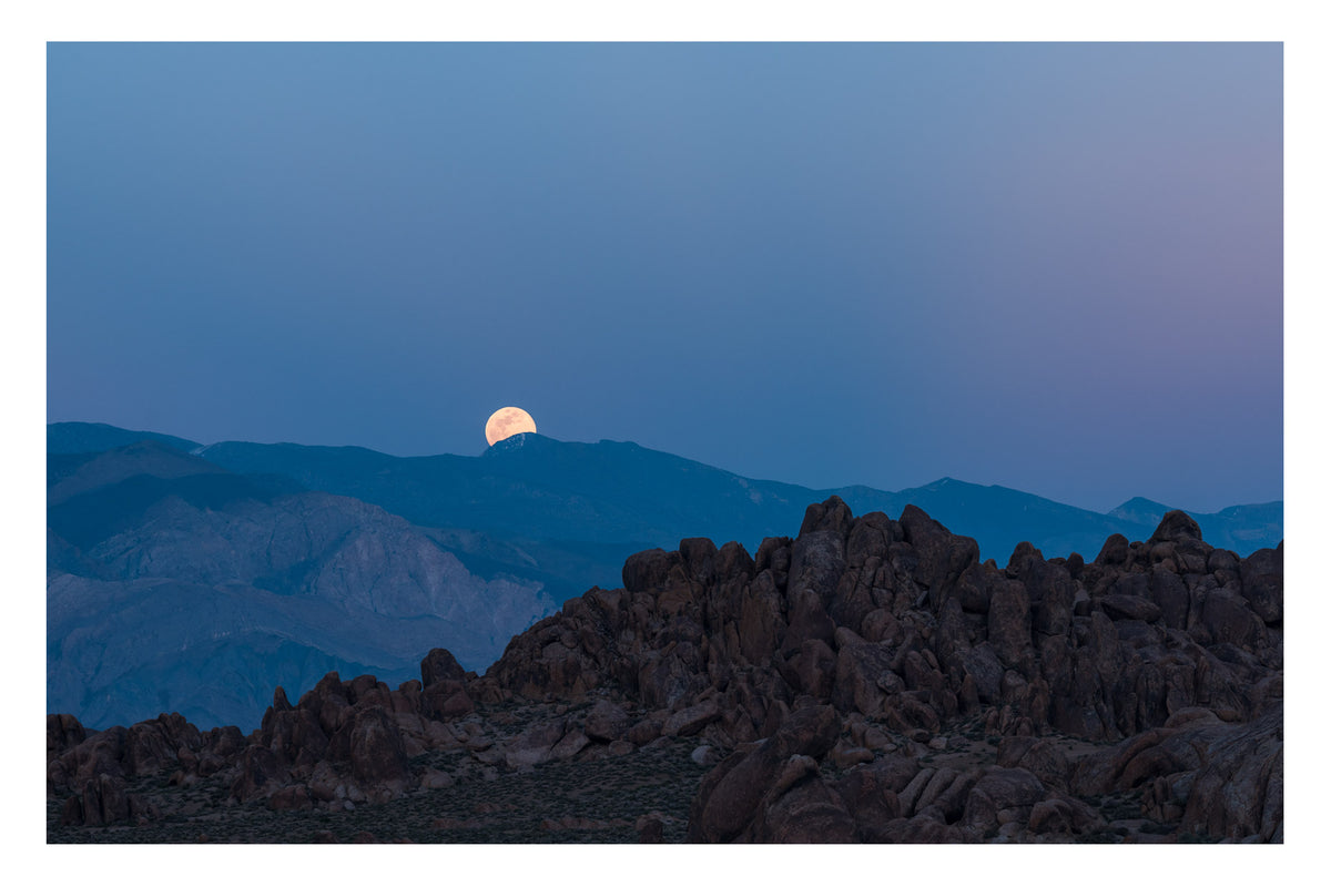 A full moon rising over the Sierra Nevada mountains above the Alabama Hills granite boulders in the blue hour, the warm peach glow of the moon contrasting against the cool graduated blue purple dusk sky. Fine art colour photographic print by Simon Davidson.