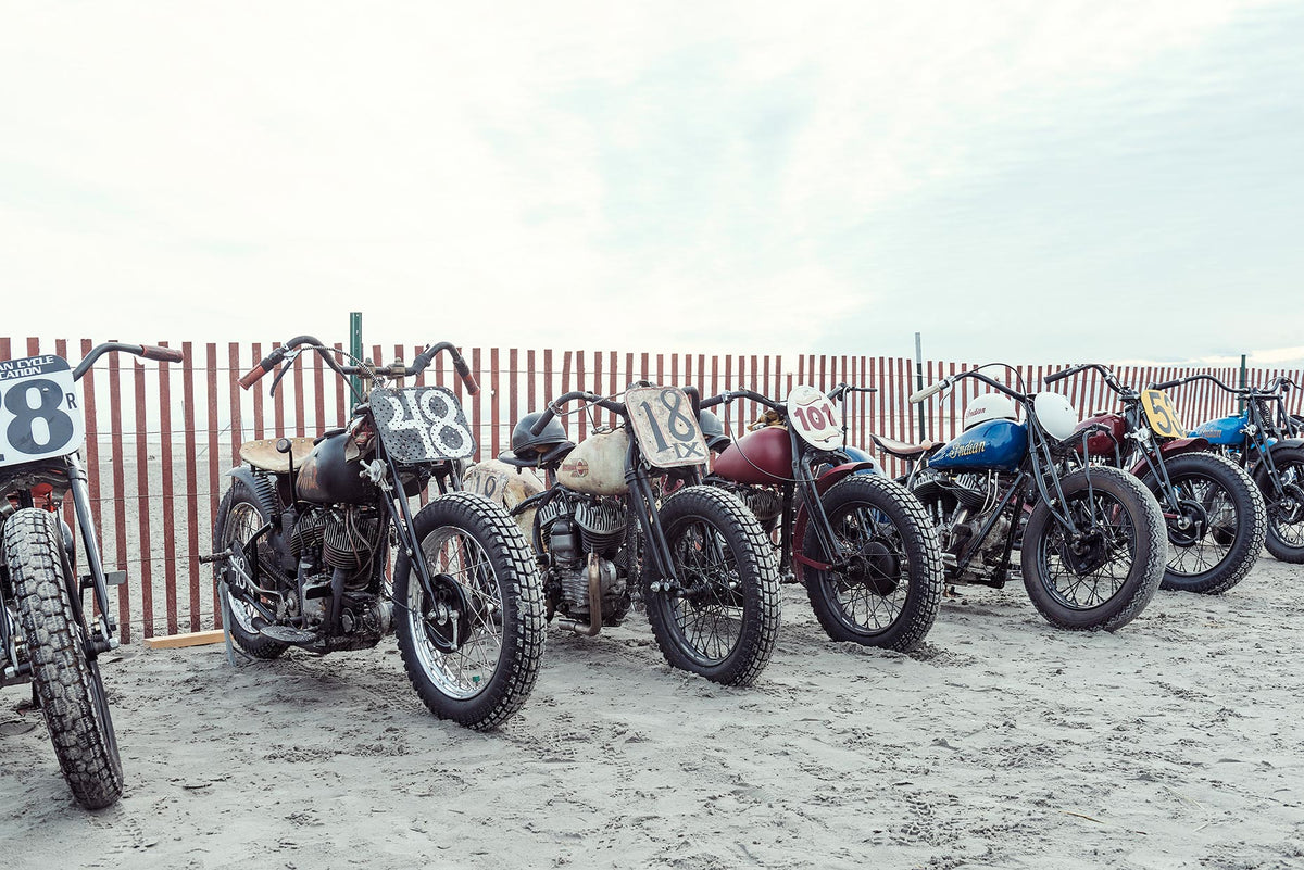 A row of pre-war vintage Harley Davidson and Indian motorcycles with hand painted race numbers parked against a beach fence in the sand, including numbers 48, 18, 101 and a blue Indian. The beach pits. Captured at the Race of Gentlemen. Fine art colour photographic print by Simon Davidson.