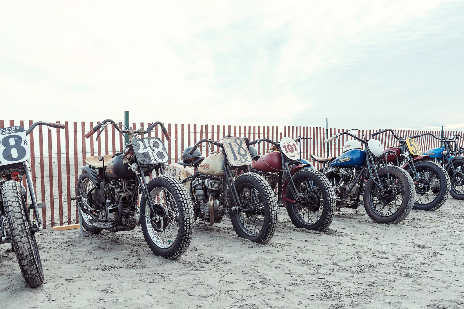 A row of pre-war vintage Harley Davidson and Indian motorcycles with hand painted race numbers parked against a beach fence in the sand, including numbers 48, 18, 101 and a blue Indian. The beach pits. Captured at the Race of Gentlemen. Fine art colour photographic print by Simon Davidson.