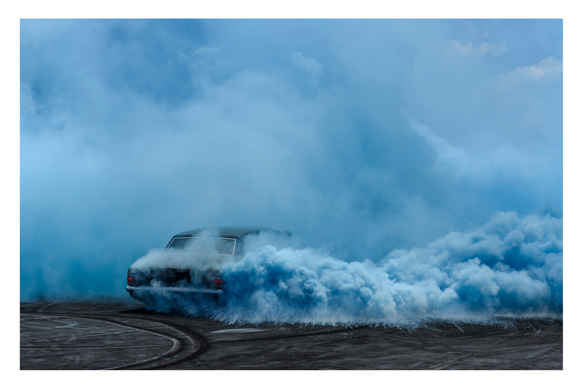 A black Holden sedan known as UCSMOKE performing a burnout, almost completely dissolved into a vast rolling cloud of deep blue tyre smoke. Fine art burnout photographic print by Simon Davidson.