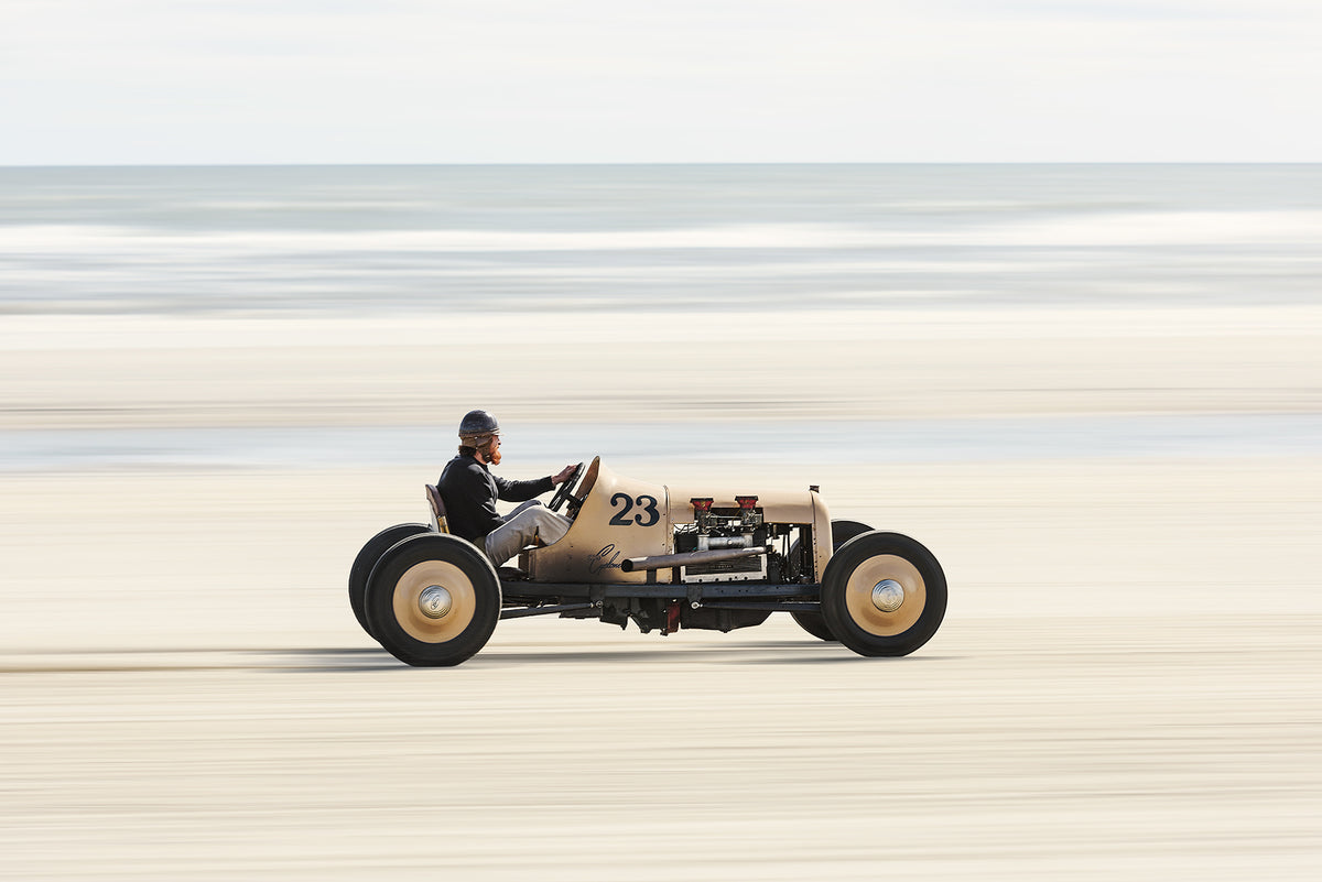 A pre-war vintage, single seat, open wheel beach racer numbered 23, cream bodied with exposed flat head engine. Captured at speed with motion blur streaking the sand and sea behind it. Captured at the Race of Gentlemen. Fine art colour photographic print by Simon Davidson.