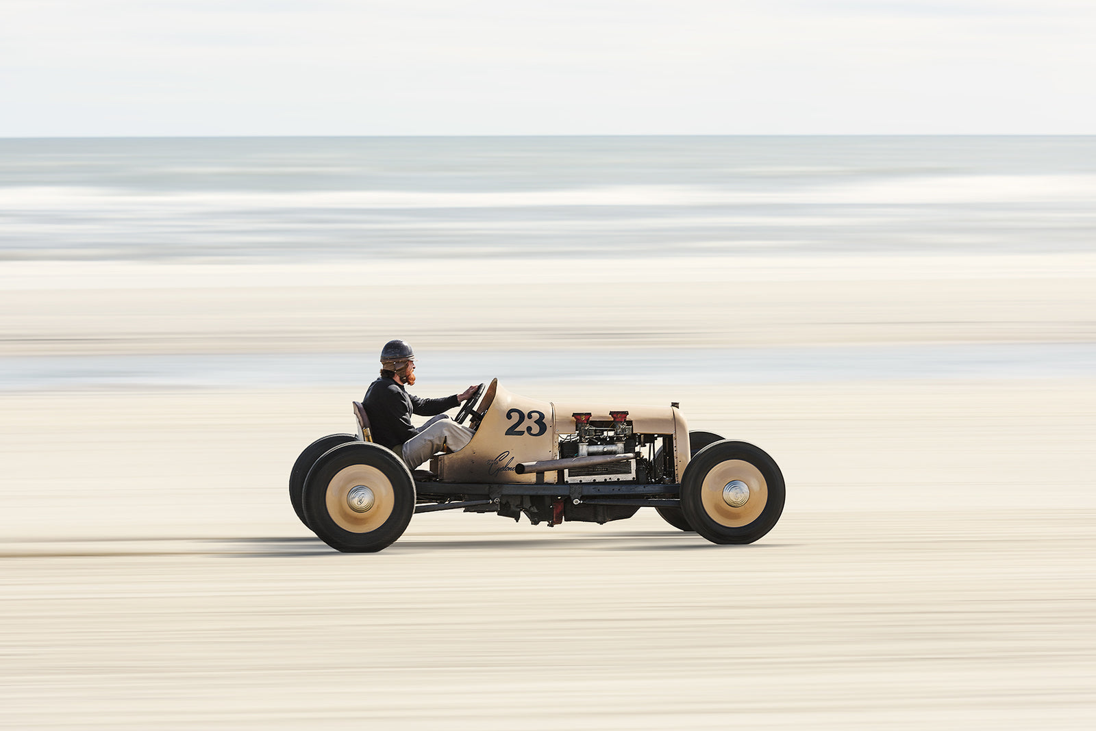 A pre-war vintage, single seat, open wheel beach racer numbered 23, cream bodied with exposed flat head engine. Captured at speed with motion blur streaking the sand and sea behind it. Captured at the Race of Gentlemen. Fine art colour photographic print by Simon Davidson.