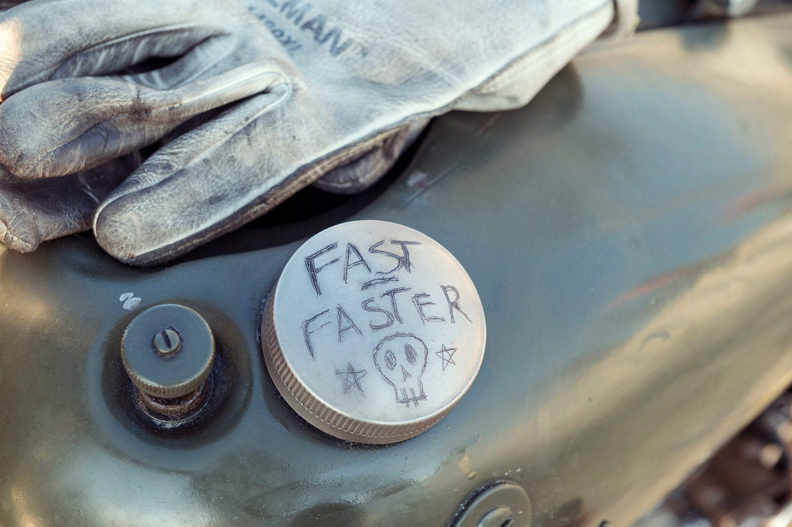 Close up of a pre-war vintage motorcycle fuel cap hand scratched with the text Fast Faster and a skull, beside a worn leather racing glove. Captured at the Race of Gentlemen. Fine art photographic print by Simon Davidson.
