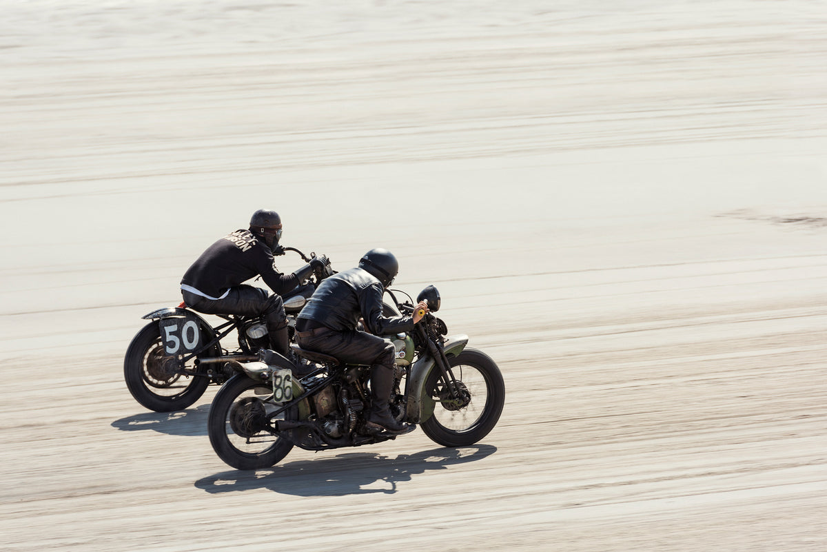 Two vintage Harley Davidson motorcycles numbered 50 and 86 racing side by side at full speed on the beach, both riders pinned low in matching black leather, the sand blurred beneath them. Captured at the Race of Gentlemen. Fine art colour photographic print by Simon Davidson.