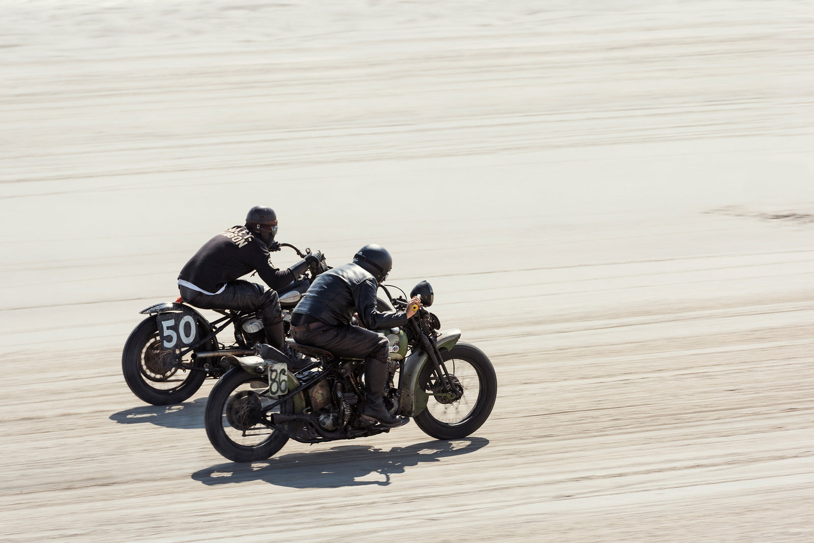 Two vintage Harley Davidson motorcycles numbered 50 and 86 racing side by side at full speed on the beach, both riders pinned low in matching black leather, the sand blurred beneath them. Captured at the Race of Gentlemen. Fine art colour photographic print by Simon Davidson.