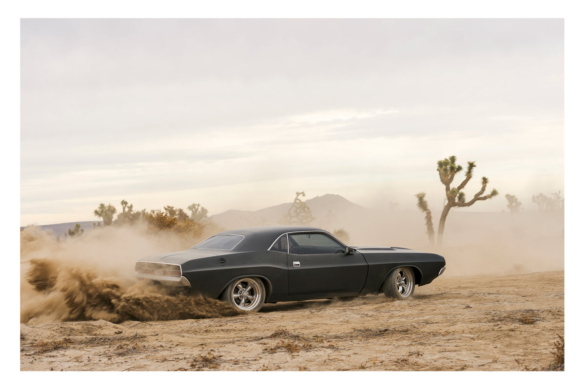 A matte black 1970 Dodge Challenger kicking up a wave of desert dirt among Joshua trees and mountains in the Mojave Desert, the dust trailing behind it like a comet's tail under a pale hazy sky. Fine art colour photographic print by Simon Davidson.