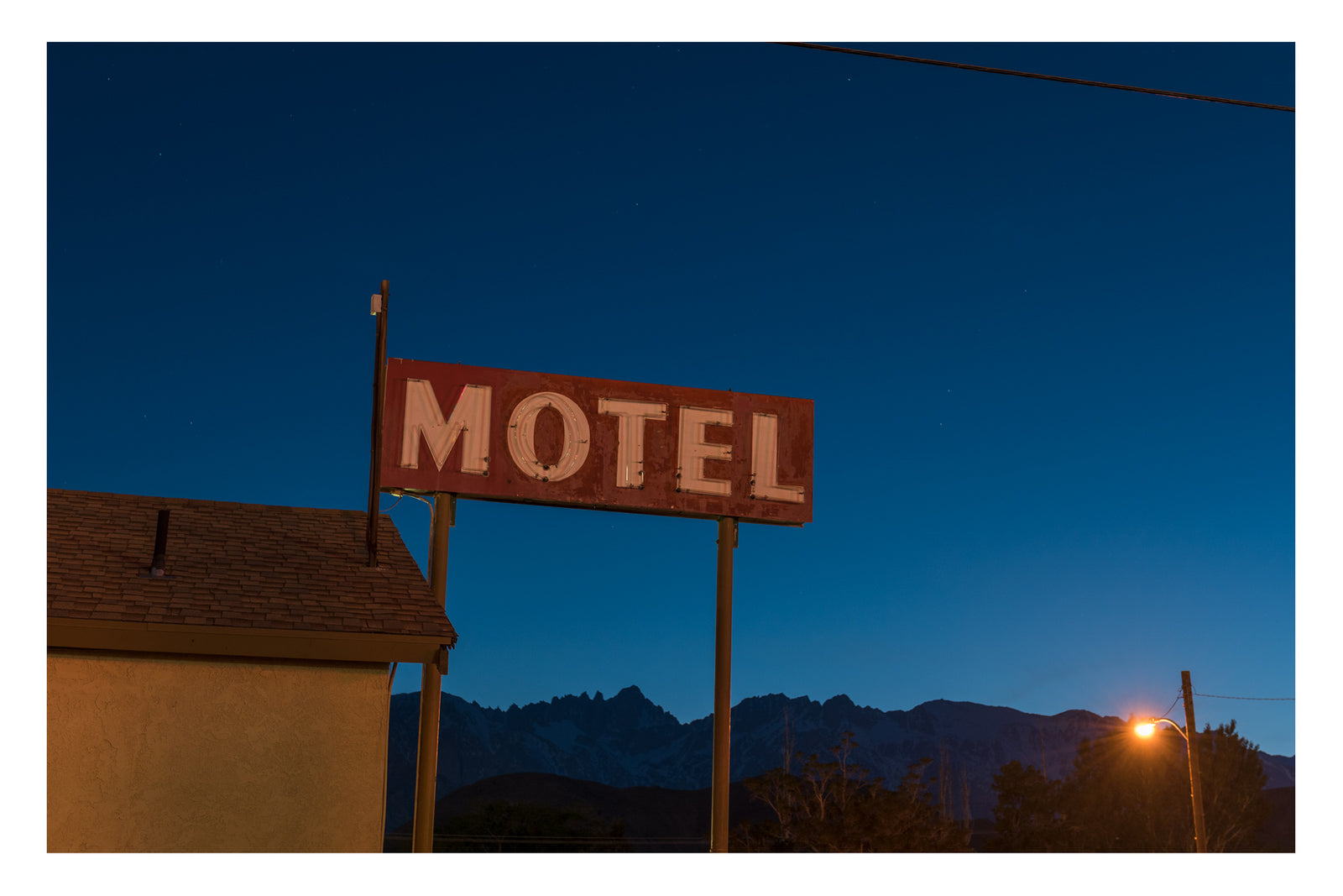 A faded vintage neon motel sign photographed at night in Lone Pine California, the jagged silhouette of the Alabama Hills and Sierra Nevada visible behind it under a deep cobalt blue sky, a single street lamp glowing warm to the lower righthand side. A palette of orange and blue. Fine art colour photographic print by Simon Davidson.