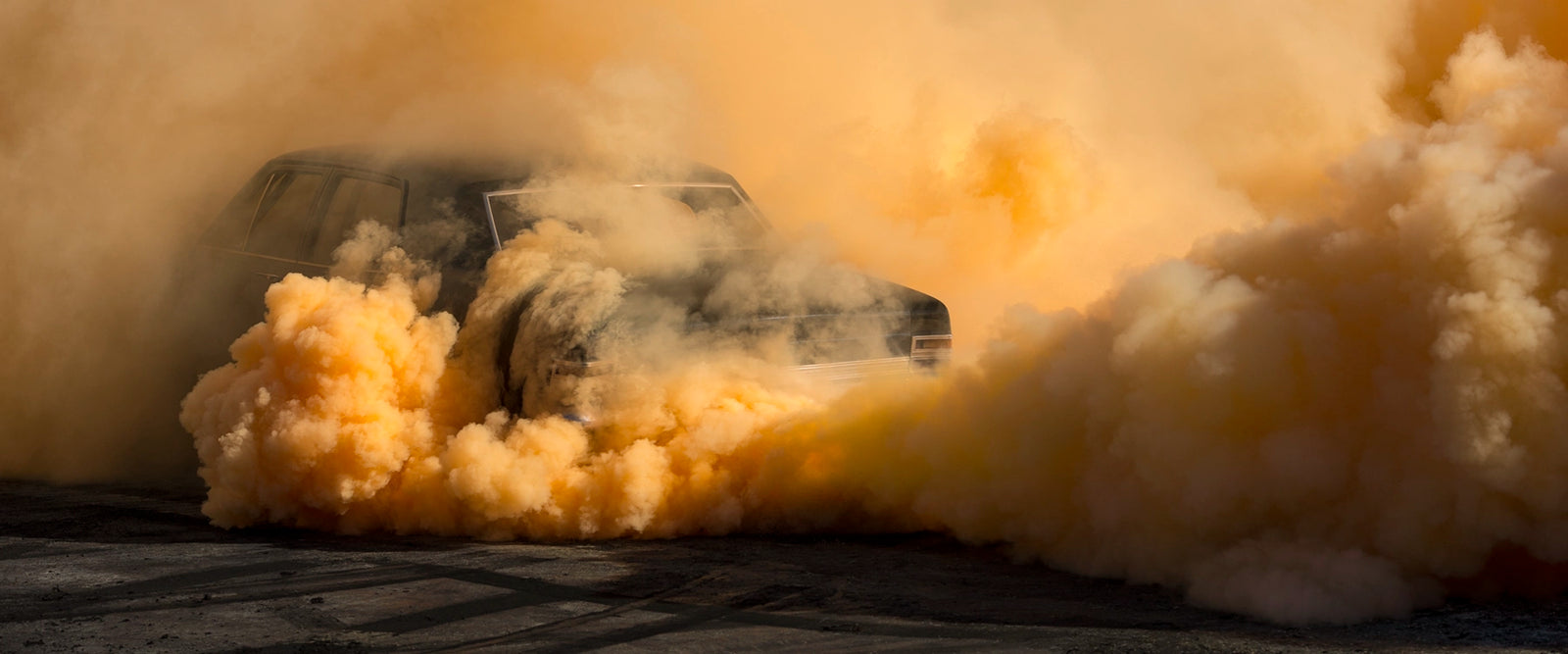 A black Holden Holden sedan is almost near consumed by a towering cloud of deep amber and orange tyre smoke that fills the entire frame, the roofline and windscreen barely visible through the churning golden haze. The warm orange smoke billows dramatically in every direction across the marked black bitumen, giving the scene an almost volcanic intensity. Fine art colour photographic print by Simon Davidson.