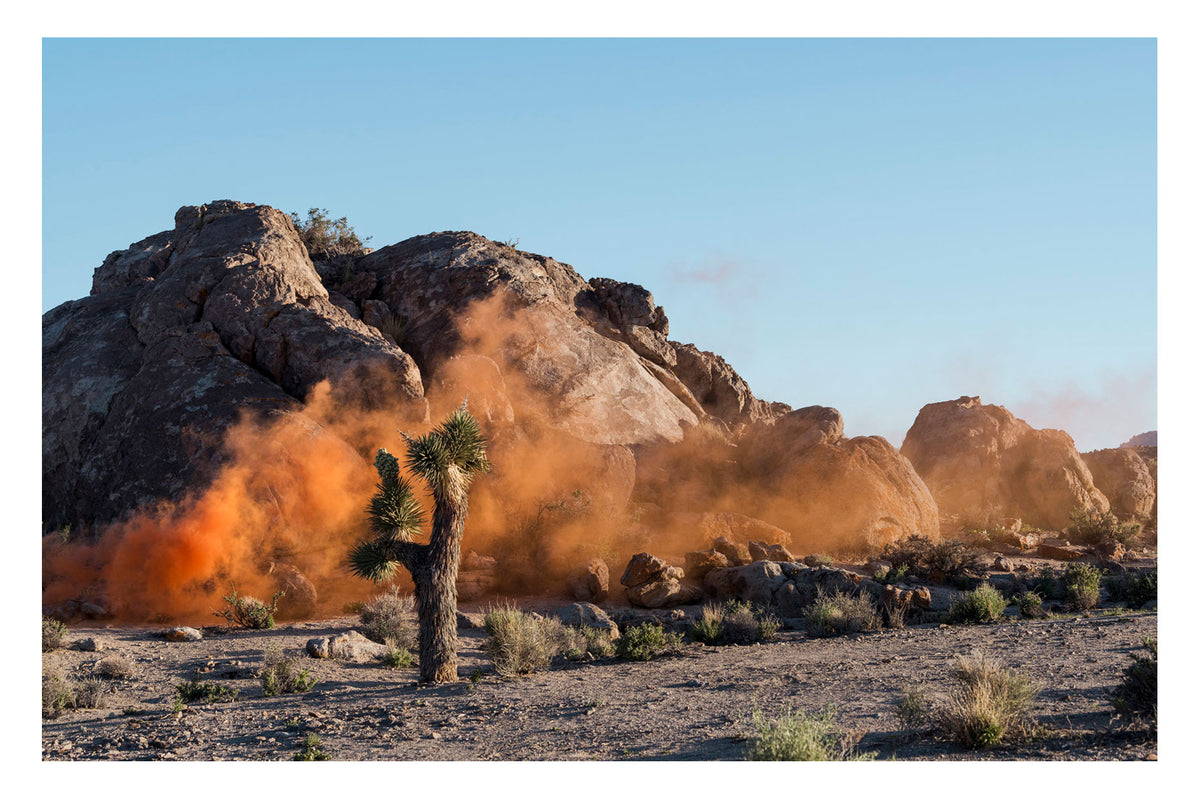 A mysterious cloud of vivid orange smoke drifts across ancient granite boulders in Joshua Tree National Park, its origin completely out of frame. A lone Joshua tree stands unmoved in the foreground. Like a film still from another world. Captured in the last rays of the sun. Shadows long. Fine art colour photographic print by Simon Davidson.