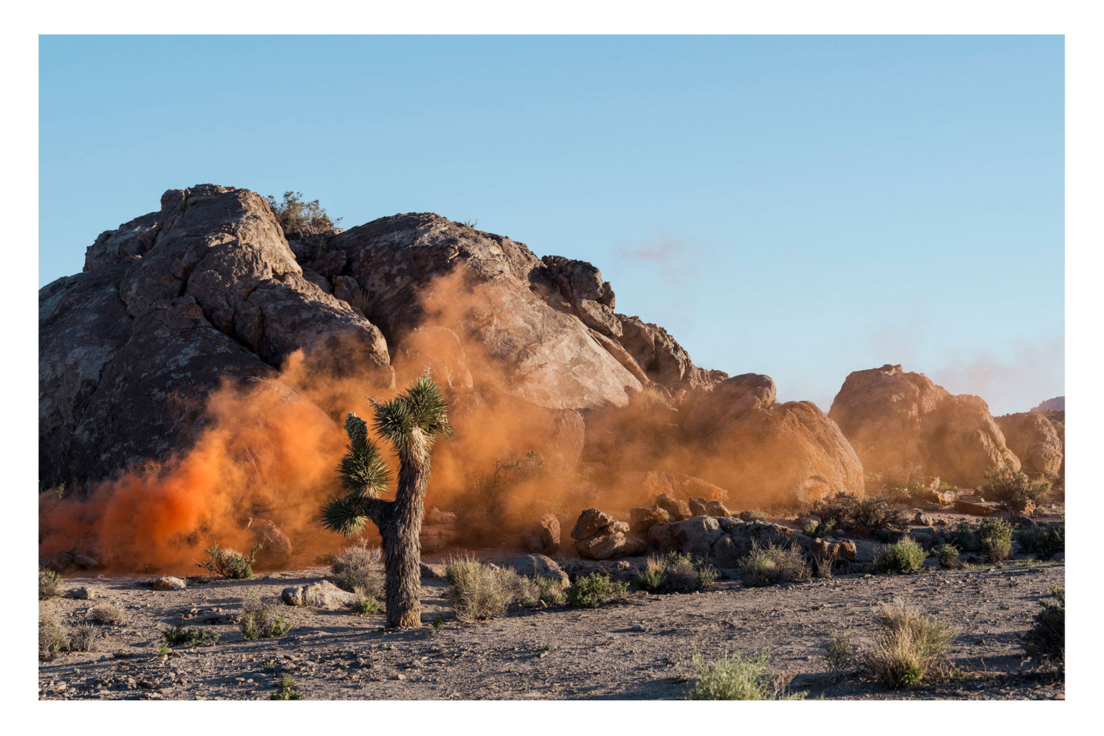 A mysterious cloud of vivid orange smoke drifts across ancient granite boulders in Joshua Tree National Park, its origin completely out of frame. A lone Joshua tree stands unmoved in the foreground. Like a film still from another world. Captured in the last rays of the sun. Shadows long. Fine art colour photographic print by Simon Davidson.
