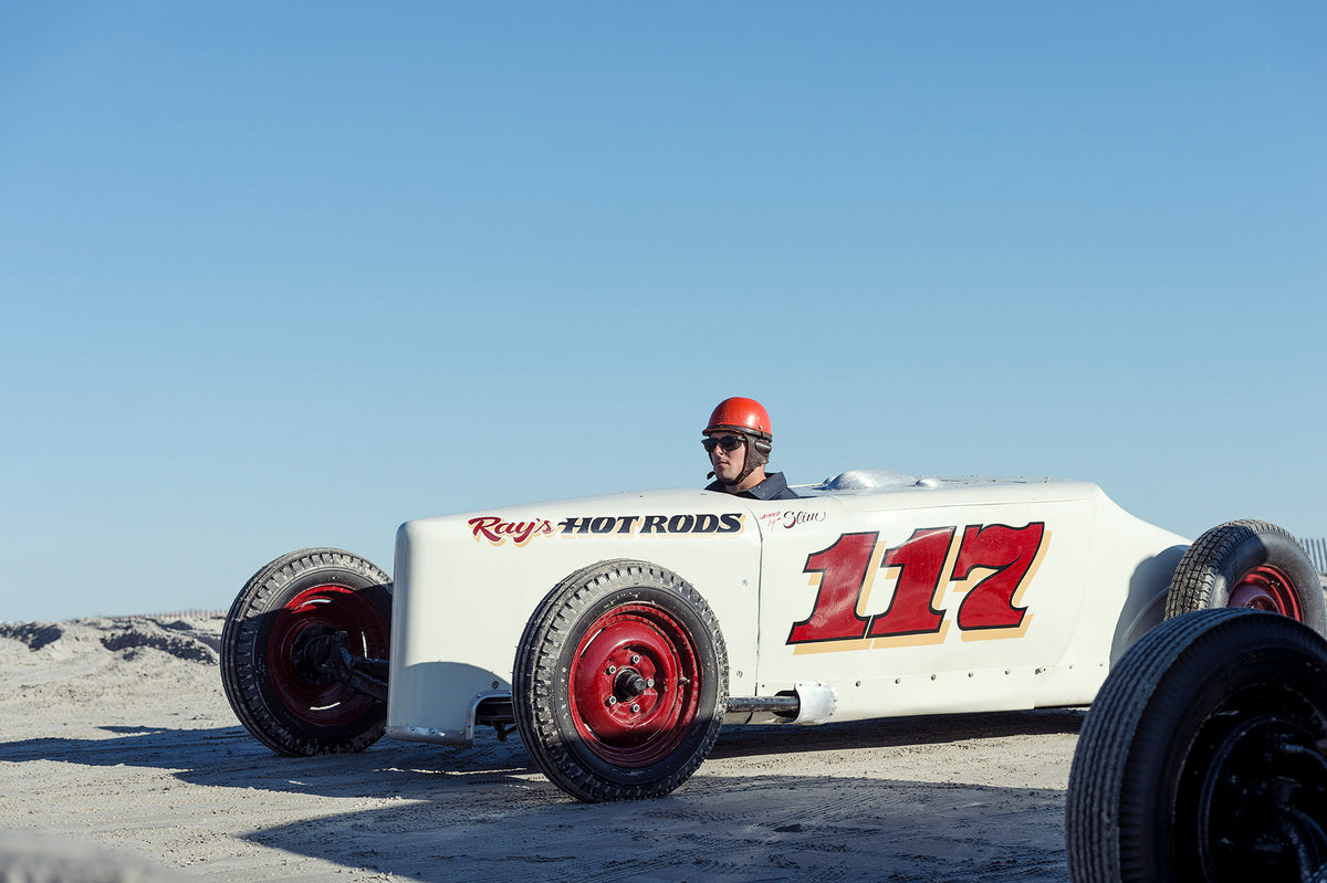 Ray's Hot Rods number 117, a white pre-war vintage beach racer with bold red race numbers and red wheels, driver wearing a red helmet and sunglasses sitting low on the sand under a clear blue sky. Captured at the Race of Gentlemen. Fine art colour photographic print by Simon Davidson.