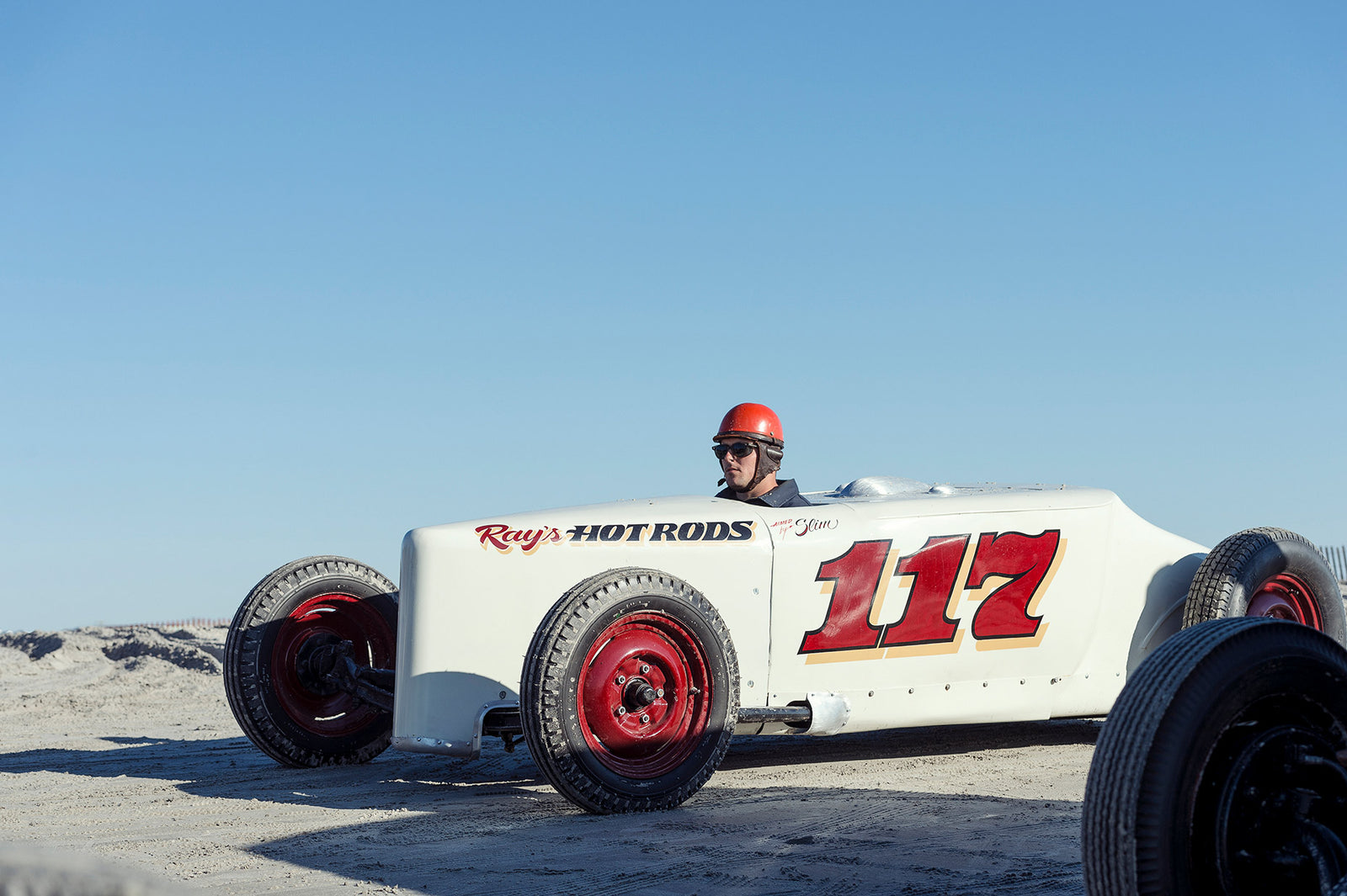 Ray's Hot Rods number 117, a white pre-war vintage beach racer with bold red race numbers and red wheels, driver wearing a red helmet and sunglasses sitting low on the sand under a clear blue sky. Captured at the Race of Gentlemen. Fine art colour photographic print by Simon Davidson.
