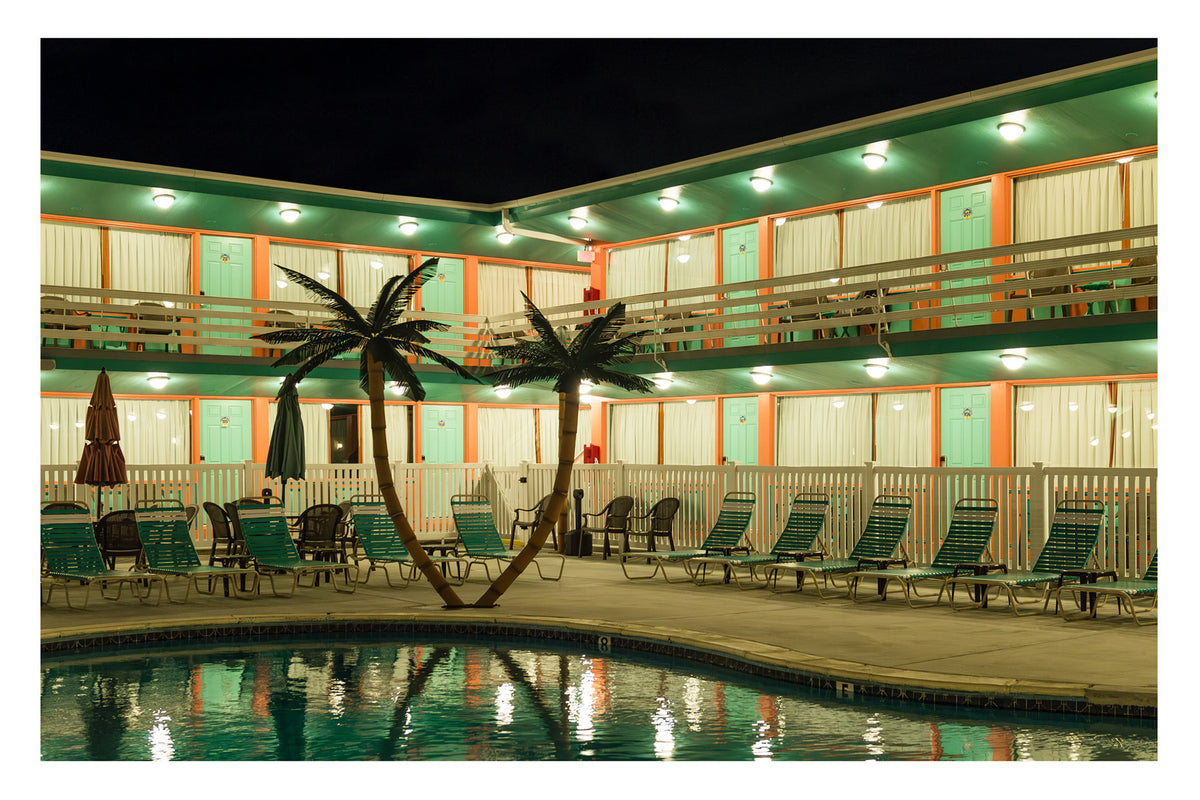 An empty 1950s mid century modern American motel courtyard photographed at night, with a glowing swimming pool in the foreground and rows of empty green sun loungers surrounding two fake palm trees. The two storey motel facade is lit. Painted in warm green and orange tones with mint coloured doors lining both levels, reflecting in the still pool water against a pitch black sky. Fine art colour photographic print by Simon Davidson.