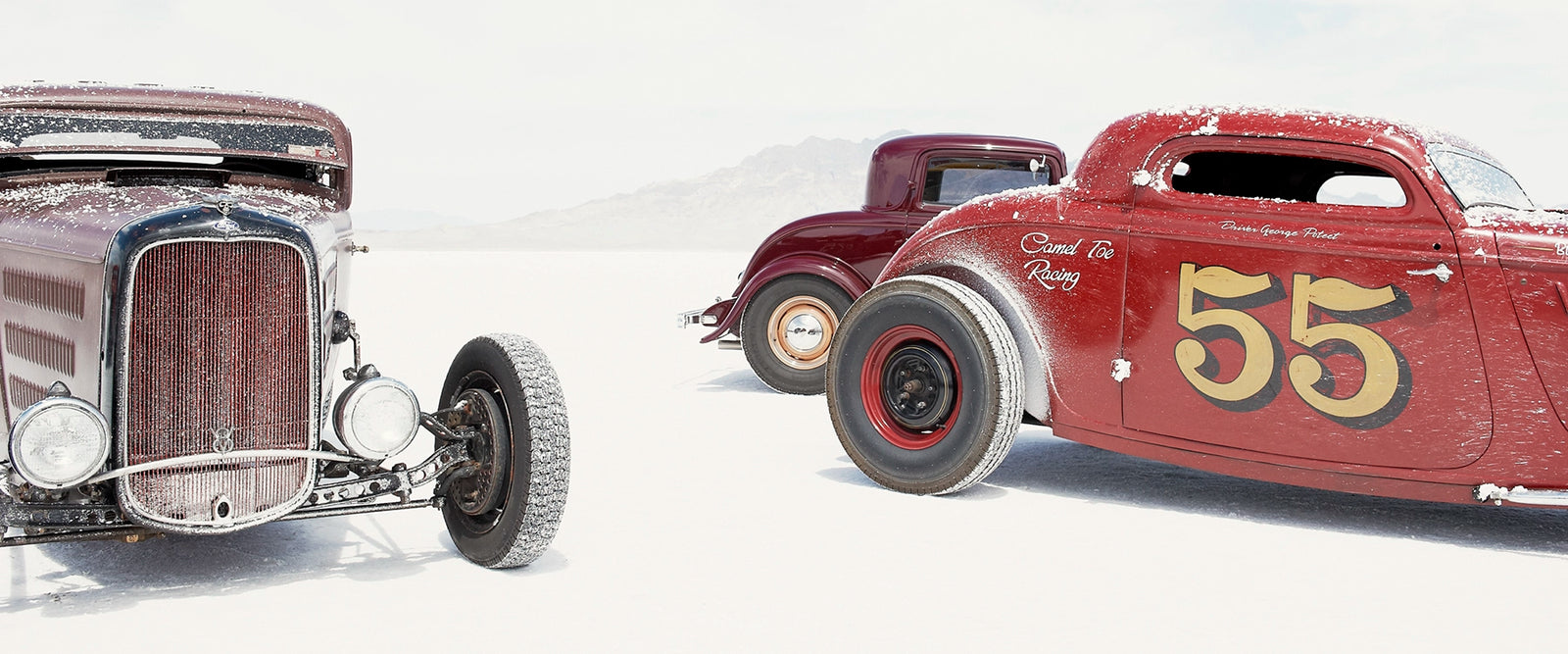 Two salt encrusted Rolling Bones hot rods sit on the blinding white surface of the Bonneville Salt Flats, their weathered bodies dusted with salt crystal residue. with a third maroon coupe visible between them in the middle distance. A pale mountain range dissolves into the white horizon behind them. Fine art colour photographic print by Simon Davidson.