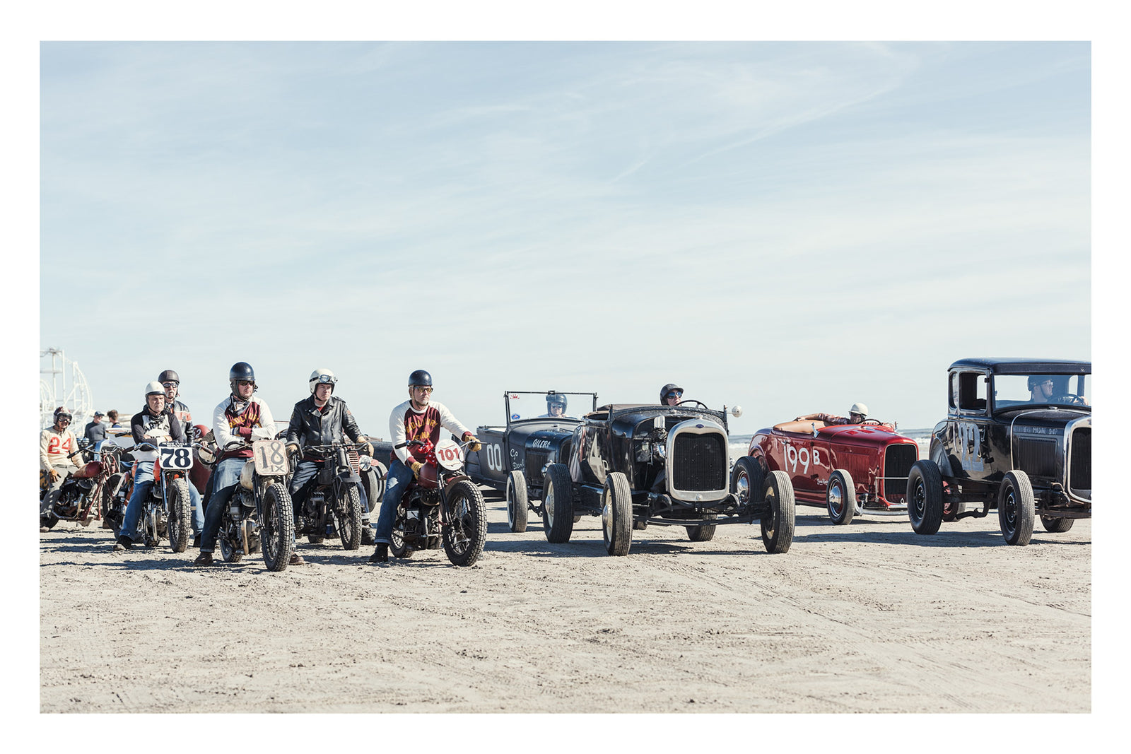 A lineup of vintage pre-war motorcycles and hot rod roadsters with their riders and drivers assembled on the beach before a race, a Ferris wheel visible in the background. Captured at the Race of Gentlemen. Fine art colour  photographic print by Simon Davidson.