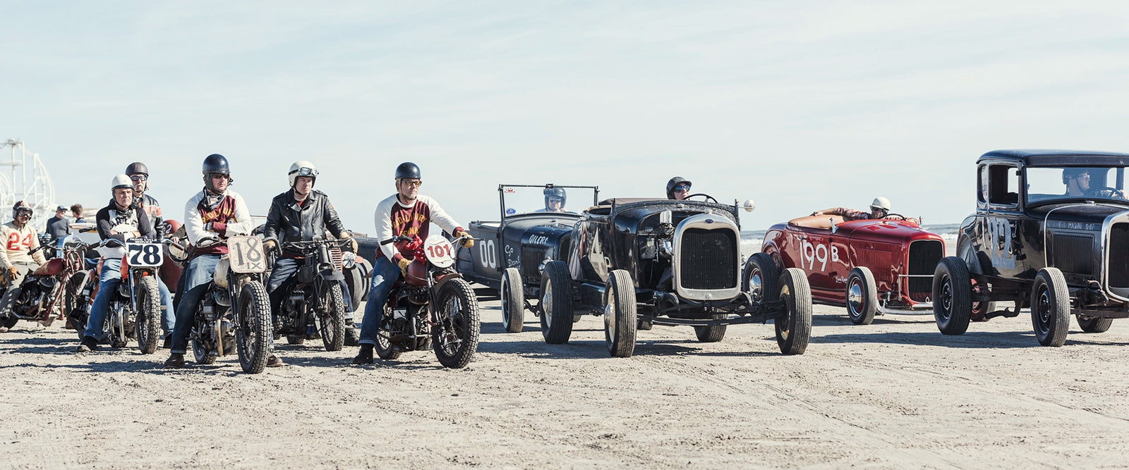 A lineup of vintage pre-war motorcycles and hot rod roadsters with their riders and drivers assembled on the beach before a race, a Ferris wheel visible in the background. Captured at the Race of Gentlemen. Fine art colour  photographic print by Simon Davidson.