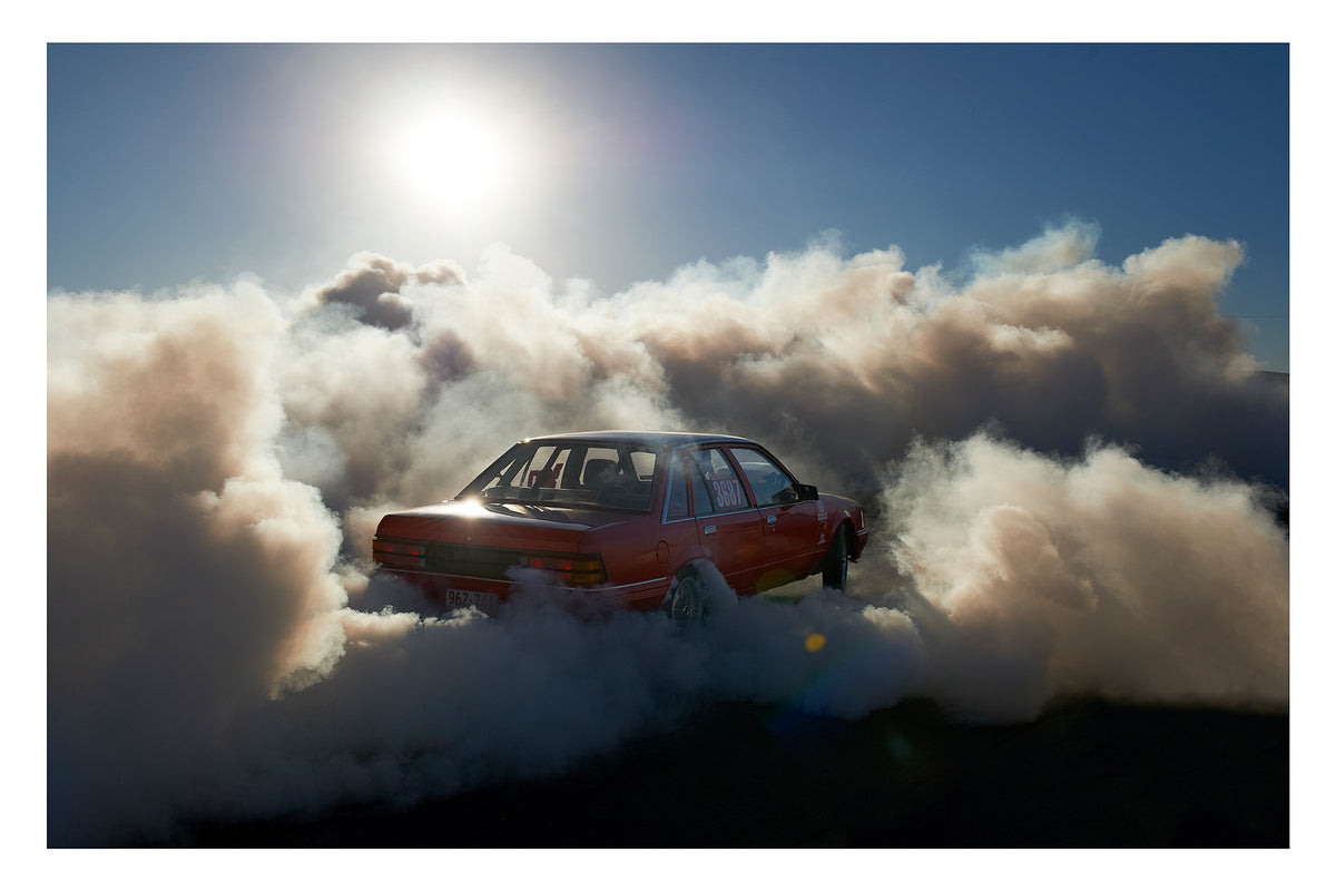 A red Holden Commodore sedan numbered 3687 performs a burnout under a blazing sun, backlit by the harsh white glare as thick white tyre smoke billows outward in every direction and glows gold where the sunlight cuts through. The stripped interior and roll cage are visible through the rear window as the car sits deep in its own cloud, the brilliant sun and deep blue central Australian sky burning above the chaos below. Fine art colour photographic print by Simon Davidson.