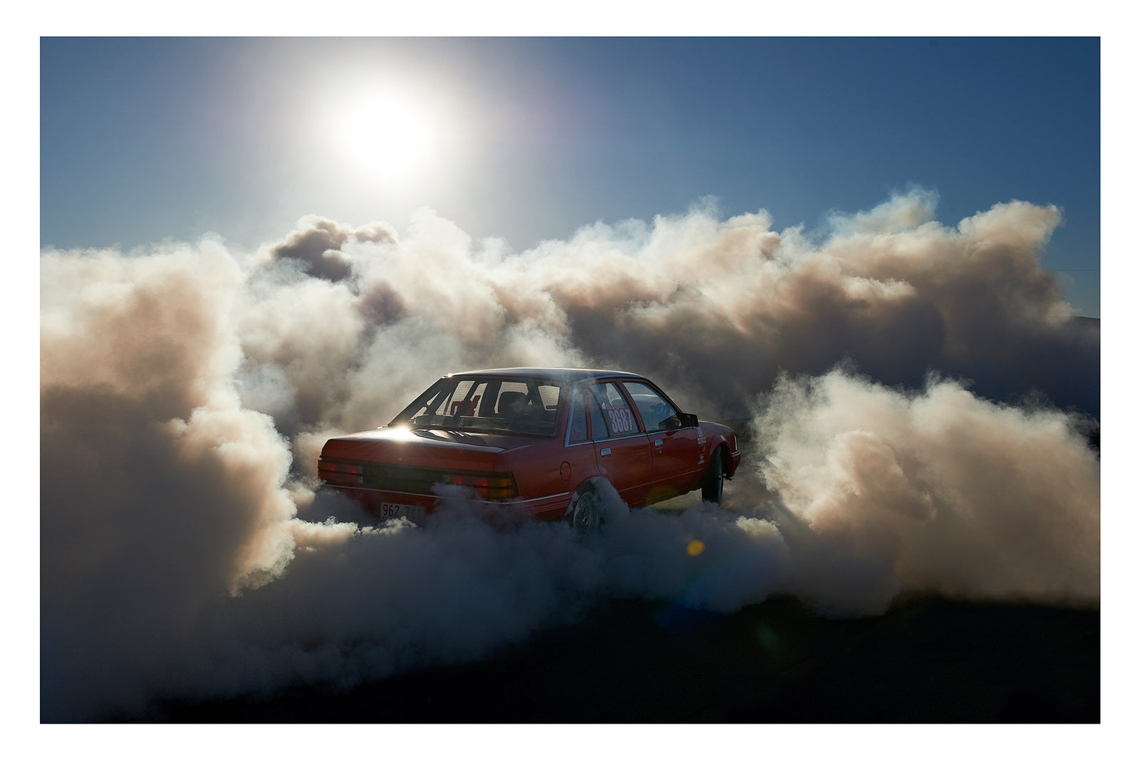 A red Holden Commodore sedan numbered 3687 performs a burnout under a blazing sun, backlit by the harsh white glare as thick white tyre smoke billows outward in every direction and glows gold where the sunlight cuts through. The stripped interior and roll cage are visible through the rear window as the car sits deep in its own cloud, the brilliant sun and deep blue central Australian sky burning above the chaos below. Fine art colour photographic print by Simon Davidson.