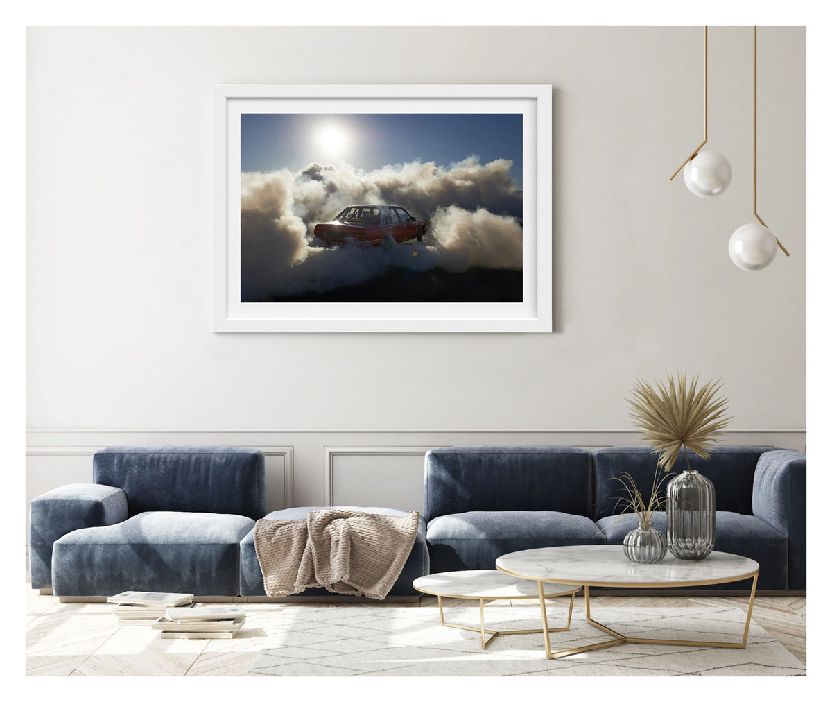 Framed in white, hanging in a contemporary space. A red Holden Commodore sedan performs a burnout under a blazing sun, backlit by the harsh white glare as thick white tyre smoke billows outward in every direction and glows gold where the sunlight cuts through. The stripped interior and roll cage are visible through the rear window as the car sits deep in its own cloud, the brilliant sun and deep blue central Australian sky burning above the chaos below. Fine art colour photographic print by Simon Davidson.