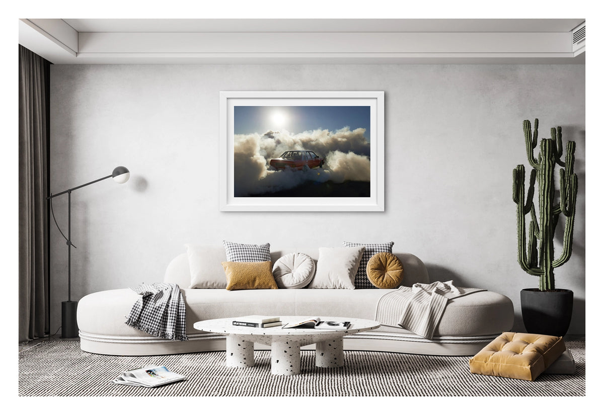 Framed in white, hanging in a contemporary space. A red Holden Commodore sedan performs a burnout under a blazing sun, backlit by the harsh white glare as thick white tyre smoke billows outward in every direction and glows gold where the sunlight cuts through. The stripped interior and roll cage are visible through the rear window as the car sits deep in its own cloud, the brilliant sun and deep blue central Australian sky burning above the chaos below. Fine art colour photographic print by Simon Davidson.