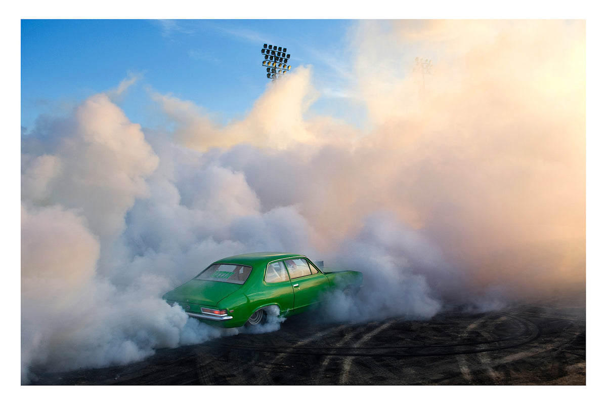 A green Holden LJ Torana sedan performs a burnout, slightly engulfed against a towering wall of white and golden tyre smoke that billows up toward a deep blue sky. Stadium flood lights pierce the haze above as the smoke catches the warm evening light, the bright green body of the car vivid against the swirling white cloud and the rubber marked black bitumen below. Fine art colour photographic print by Simon Davidson.