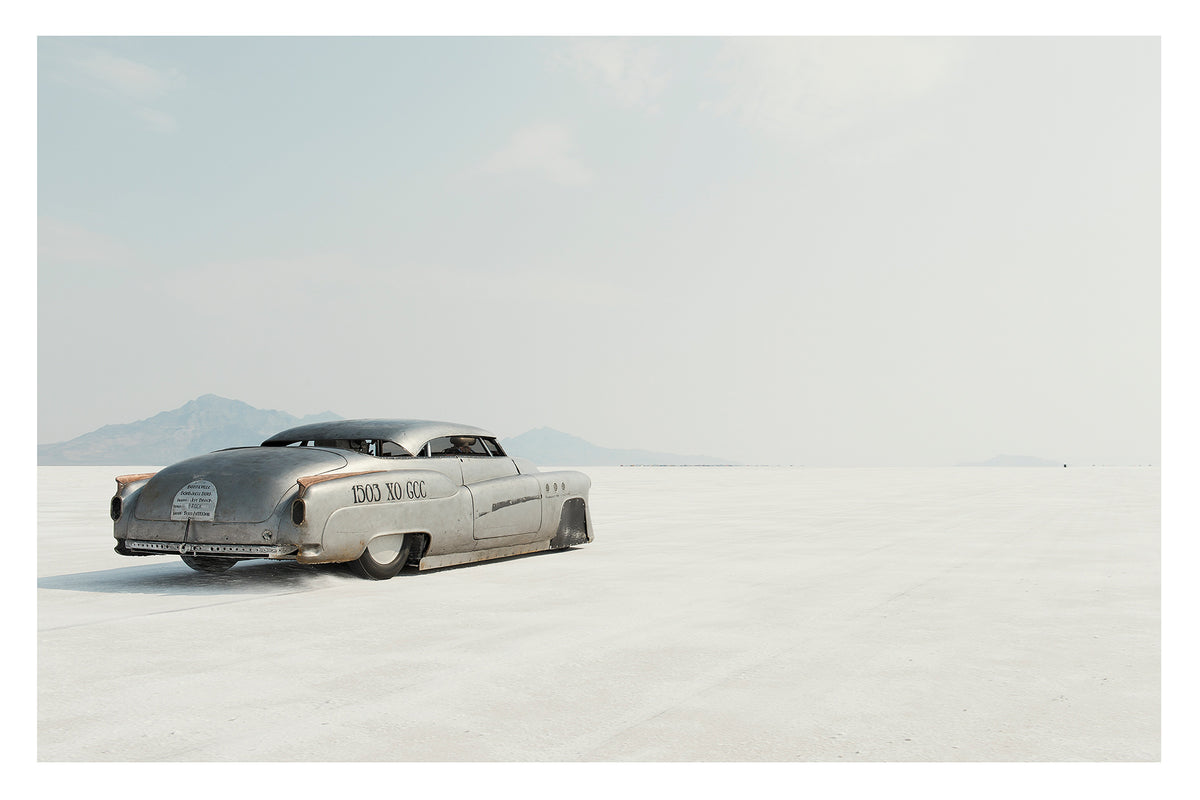 A rear three quarter view of a bare metal 1950s Buick landspeed race car sits low as it leaves the startline of the Bonneville Salt Flats, its chopped and channelled body heading towards nothingness. The bare metal bodywork and sculptural streamlined shape give the car an otherworldly, timeless presence against the vast white emptiness surrounding it. Fine art colour photographic print by Simon Davidson. 