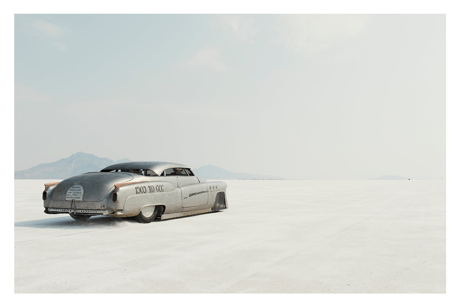 A rear three quarter view of a bare metal 1950s Buick landspeed race car sits low as it leaves the startline of the Bonneville Salt Flats, its chopped and channelled body heading towards nothingness. The bare metal bodywork and sculptural streamlined shape give the car an otherworldly, timeless presence against the vast white emptiness surrounding it. Fine art colour photographic print by Simon Davidson. 
