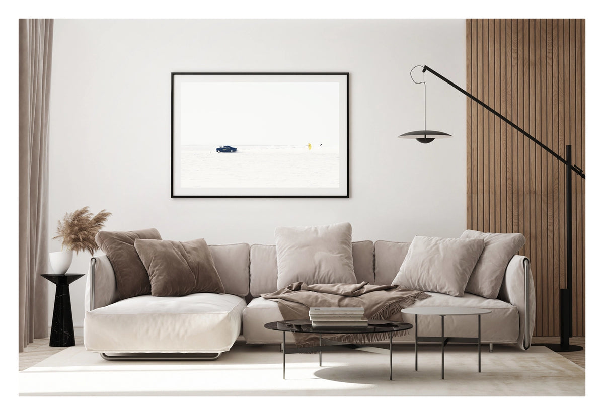 Framed in black, shown in a contemporary living space. A dark blue 1940s coupe landspeed race car numbered releases a bright yellow parachute to slow down after a high speed run across the Bonneville Salt Flats, the chute billowing open behind it as a trail of disturbed salt dust follows in its wake. The car and chute sit as tiny vivid forms against the vast featureless white of the salt and sky, the scene almost entirely bleached of colour. Fine art colour photographic print by Simon Davidson.
