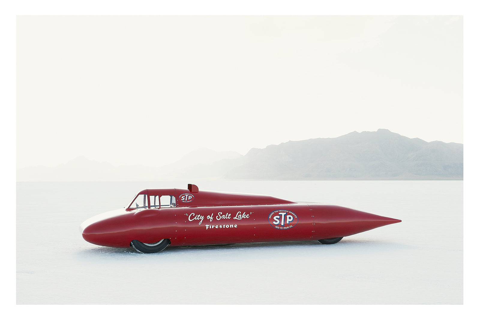 A red vintage streamliner named City of Salt Lake and sponsored by STP and Firestone sits on the Bonneville Salt Flats, its long streamlined shaped body tapering to a sharp nose point with a small raised cockpit canopy above. The elegant mid century streamliner form glows deep red against the bleached white salt and pale hazy mountains dissolving into the horizon behind it. Fine art colour photographic print by Simon Davidson.