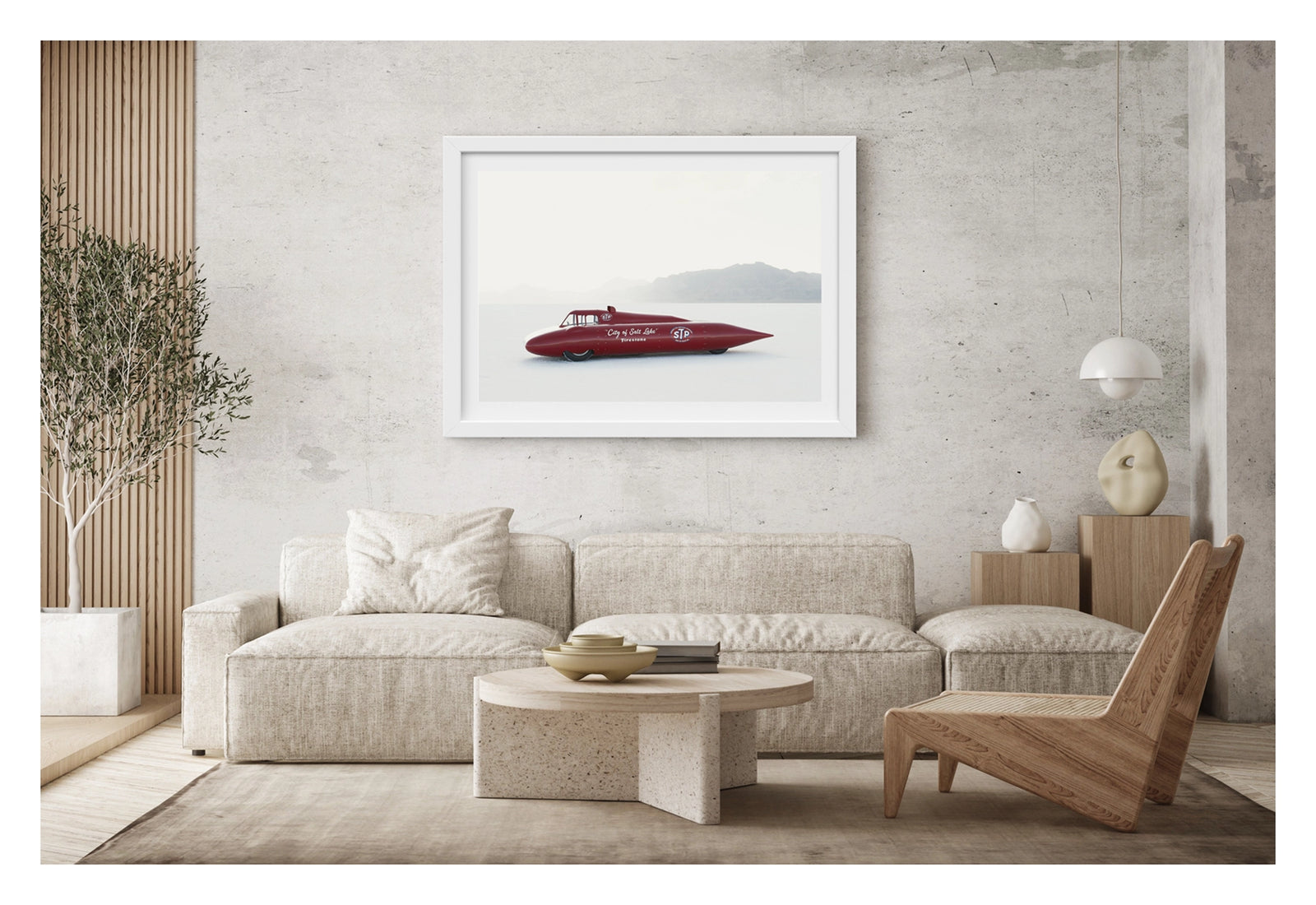 Framed in white, hanging in a contemporary living space. A red vintage streamliner named City of Salt Lake and sponsored by STP and Firestone sits on the Bonneville Salt Flats, its long streamlined shaped body tapering to a sharp nose point with a small raised cockpit canopy above. The elegant mid century streamliner form glows deep red against the bleached white salt and pale hazy mountains dissolving into the horizon behind it. Fine art colour photographic print by Simon Davidson.
