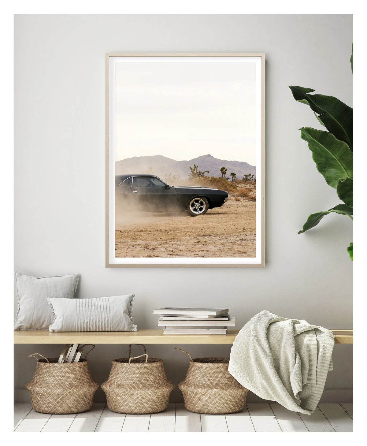 In a portrait orientation. Framed in timber. A matte black first generation Dodge Challenger powers through a desert dirt surface, kicking up a low trail of dust from the rear wheels. Joshua trees and purple hue mountain ranges stretch across the horizon under a pale white sky, with the car's polished five spoke wheels and fastback roofline sharp against the hazy desert landscape. Shown in a contemporary space. Fine art colour photographic print by Simon Davidson.