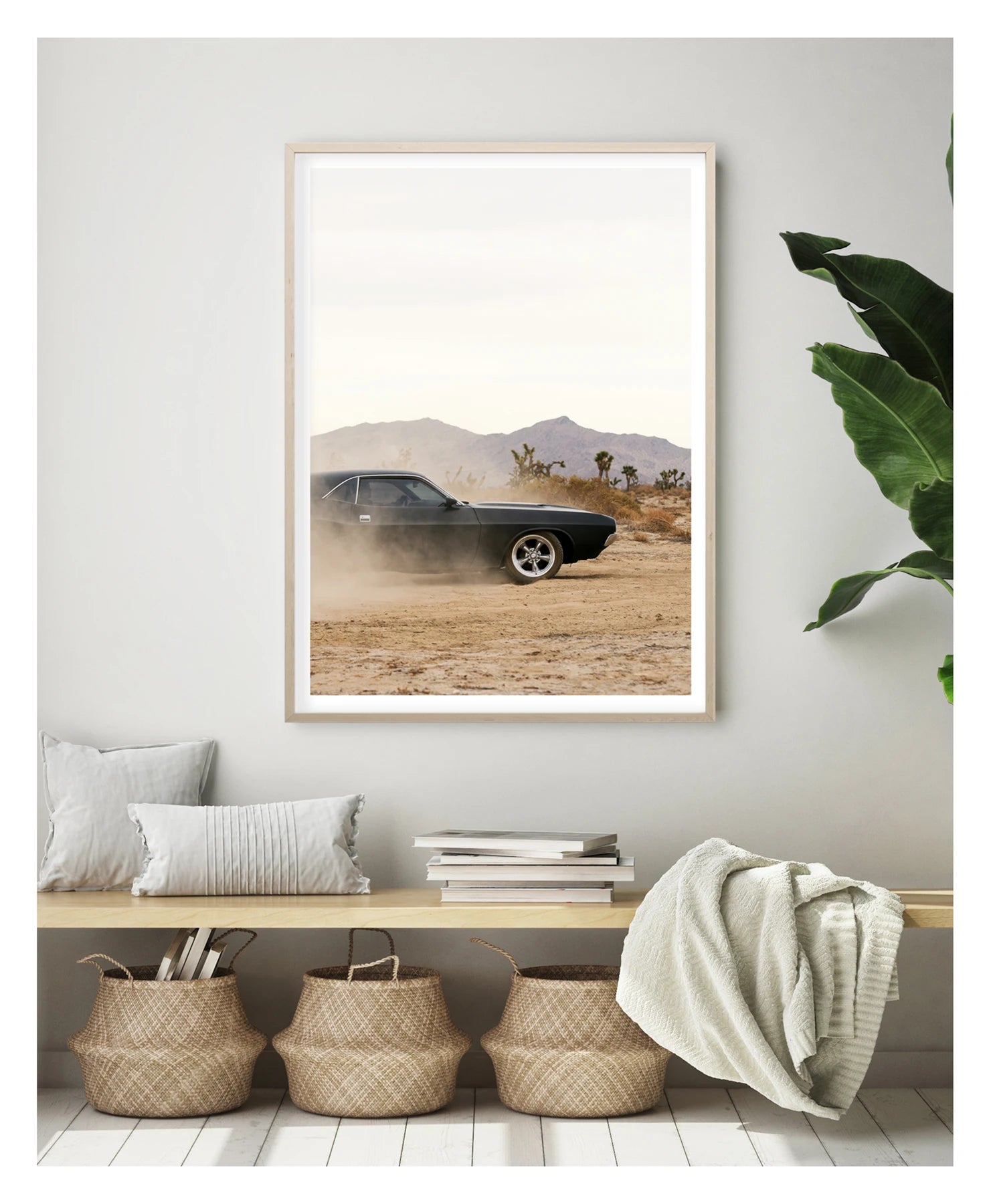 In a portrait orientation. Framed in timber. A matte black first generation Dodge Challenger powers through a desert dirt surface, kicking up a low trail of dust from the rear wheels. Joshua trees and purple hue mountain ranges stretch across the horizon under a pale white sky, with the car's polished five spoke wheels and fastback roofline sharp against the hazy desert landscape. Shown in a contemporary space. Fine art colour photographic print by Simon Davidson.