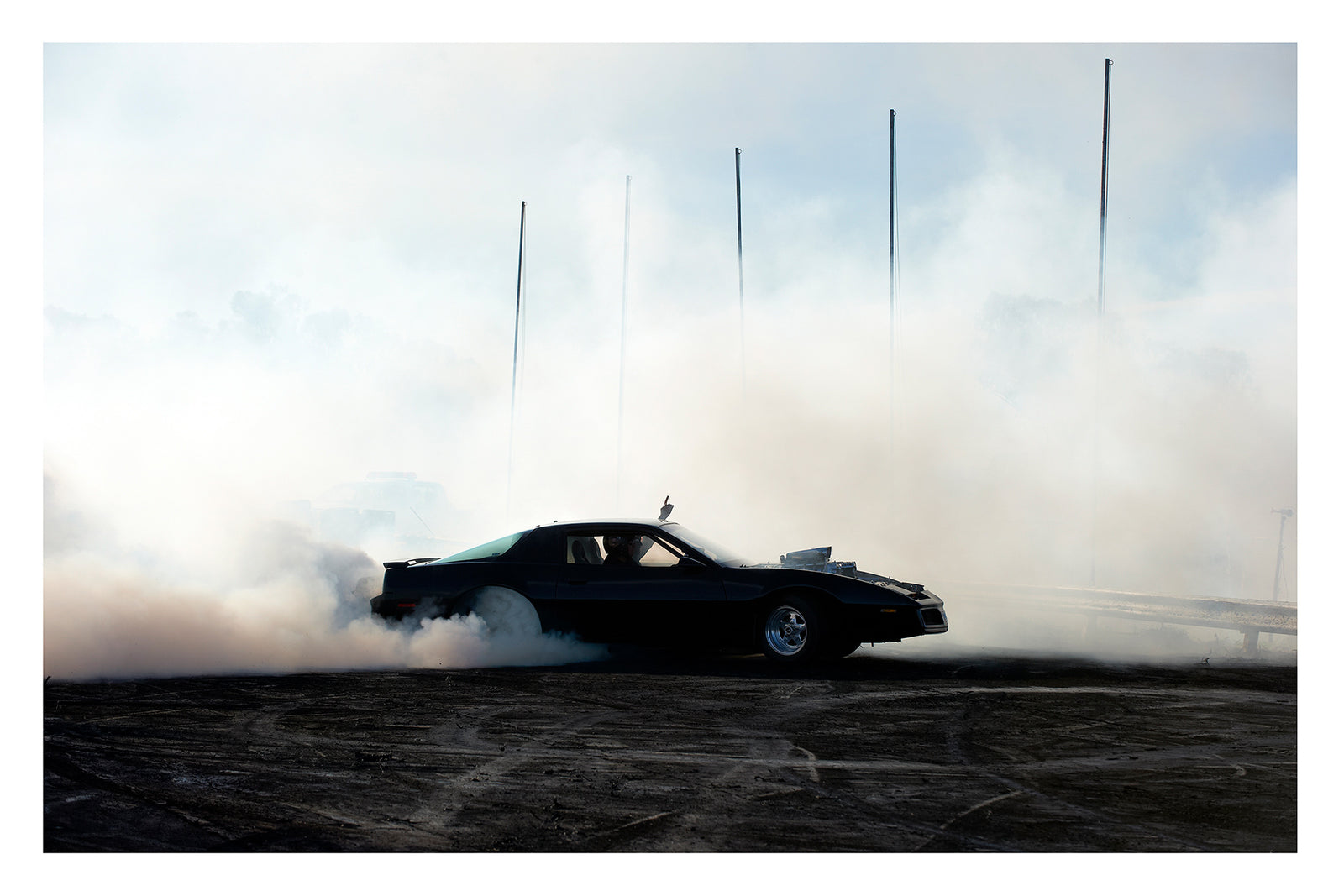 A black Pontiac Firebird executes a burnout on rubber marked black bitumen, white tyre smoke rolling low across the ground and rising behind the car as thin marker poles pierce the haze against a pale overcast sky. The dark silhouette of the car sits in sharp contrast to the surrounding smoke, a single finger raised through the open window above the roofline. Fine art colour photographic print by Simon Davidson.