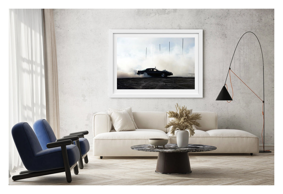 Framed in white, hanging in a contemporary living space. We see a black Pontiac Firebird executes a burnout on rubber marked black bitumen, white tyre smoke rolling low across the ground and rising behind the car as thin marker poles pierce the haze against a pale overcast sky. The dark silhouette of the car sits in sharp contrast to the surrounding smoke, a single finger raised through the open window above the roofline. Fine art colour photographic print by Simon Davidson.